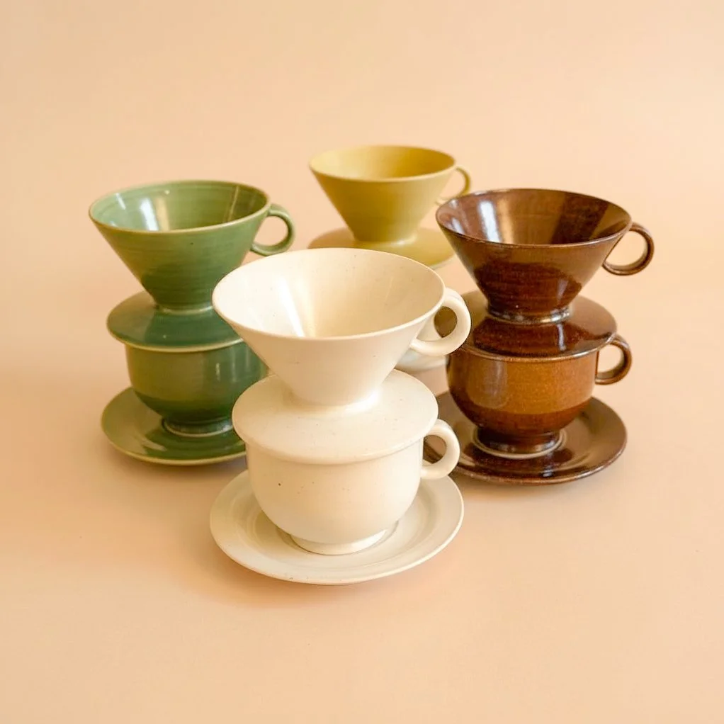 Ceramic pour-over coffee drippers in Rituals colors with matching mugs and saucers, styled on peach background for the Pooterie Studio collection.