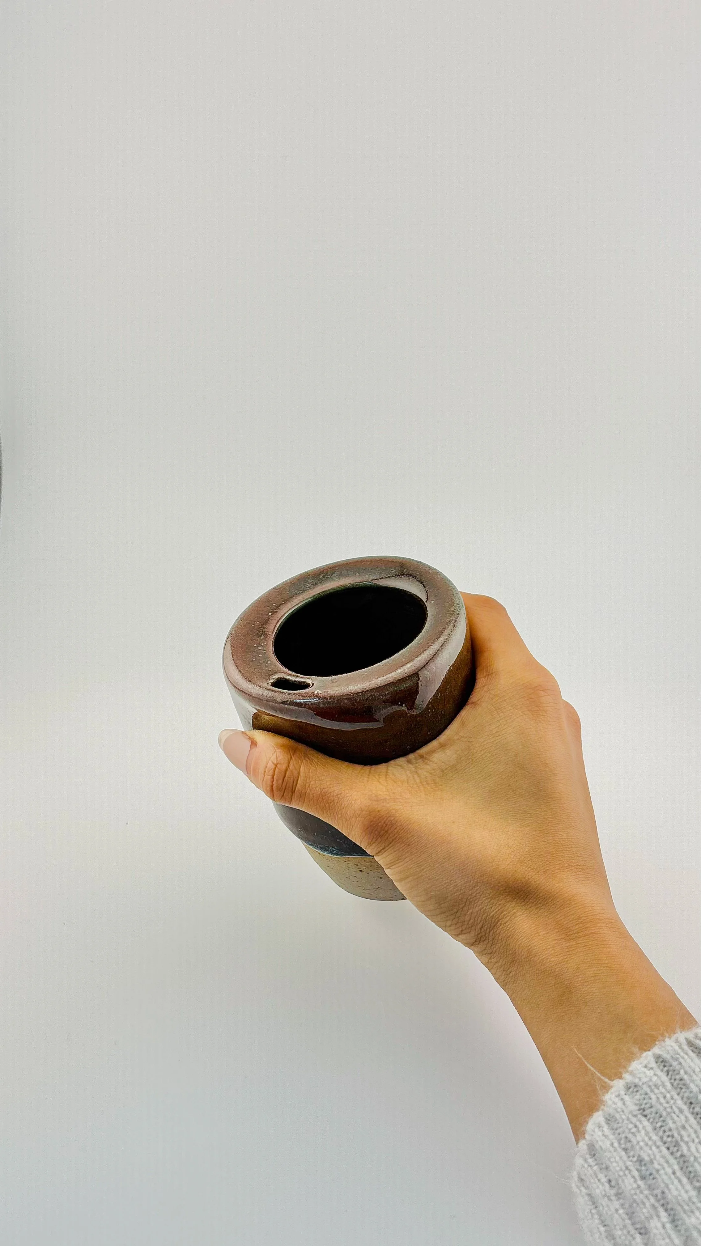 Handmade ceramic travel cup held in hand showing wide angled flange rim, deep burgundy reduction glaze with unglazed stoneware base, wheel-thrown pottery.JPG
