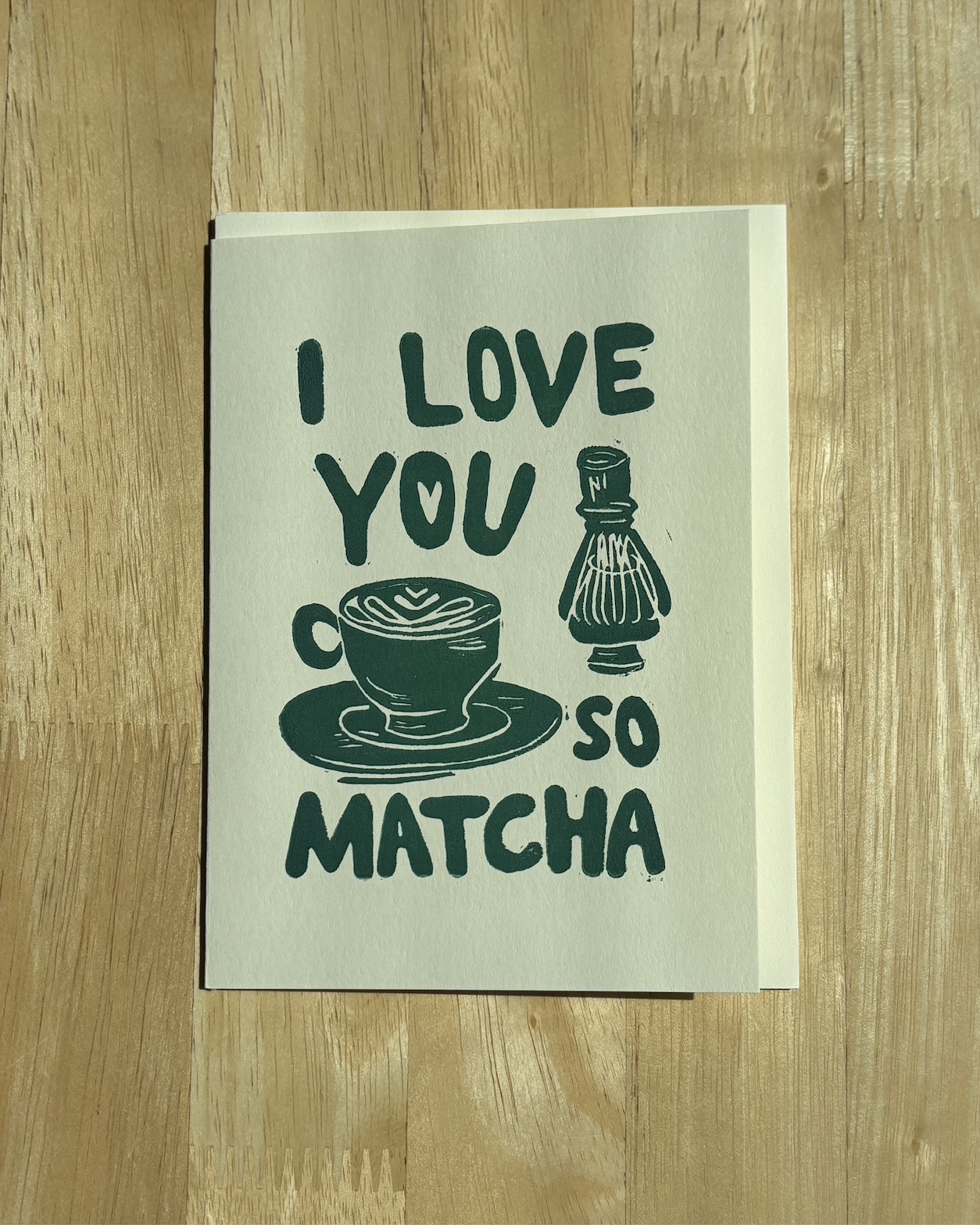 "I Love You So Matcha" linocut print card in forest green with a matcha latte and chasen design, shown with a white envelope on a warm wood table..png