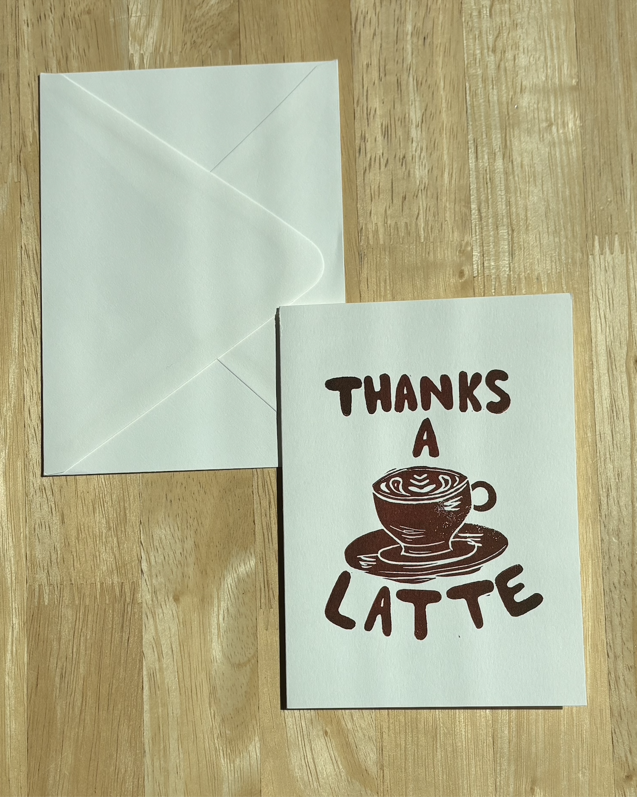 "Thanks a Latte" hand-printed linocut card in warm brown ink with a latte art illustration, paired with a white envelope on a wood surface..png