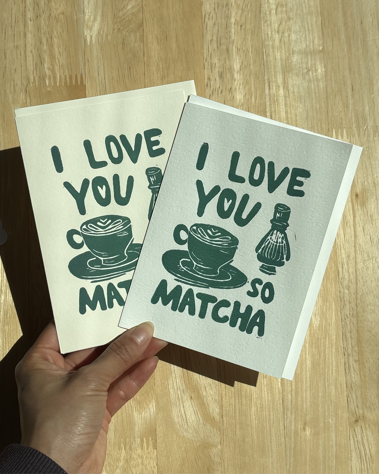 Two "I Love You So Matcha" linocut greeting cards in forest green held in hand, showing the matcha latte and bamboo chasen design side by side..png