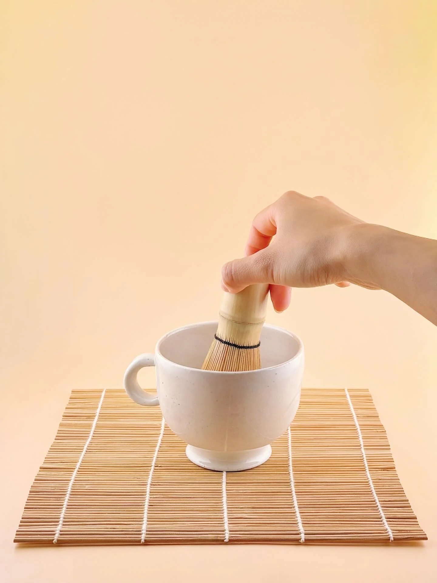 Hand whisking matcha inside white ceramic matcha mug on bamboo mat.