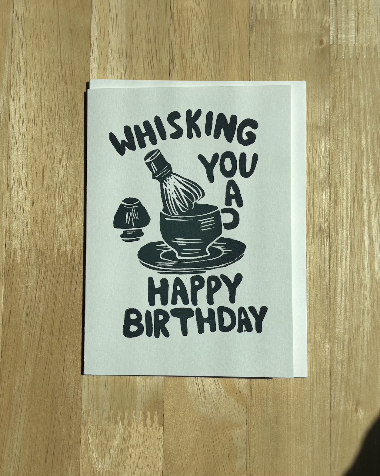 "Whisking You a Happy Birthday" hand-carved linocut card in forest green with a matcha bowl and chasen illustration, displayed with a white envelope on a wood table..png