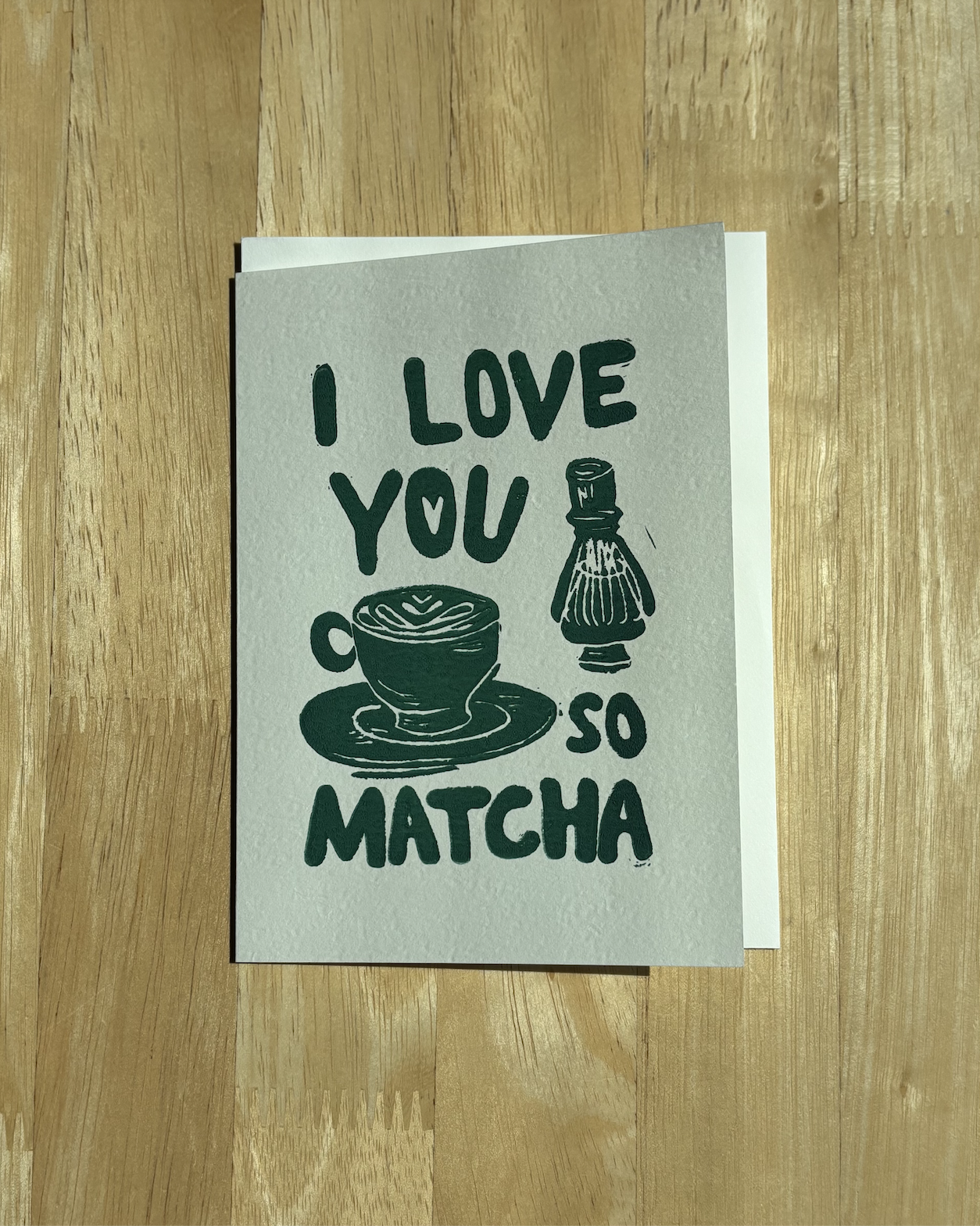 "I Love You So Matcha" hand-printed linocut card in deep green featuring a matcha latte and bamboo chasen, paired with a white envelope on a wood background..png