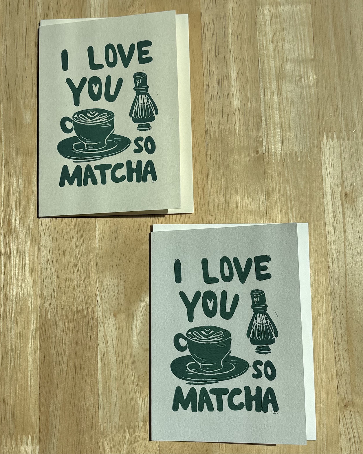 Two "I Love You So Matcha" hand-printed linocut cards in deep green stacked together, displaying the matcha latte and chasen illustration on a wood table..png
