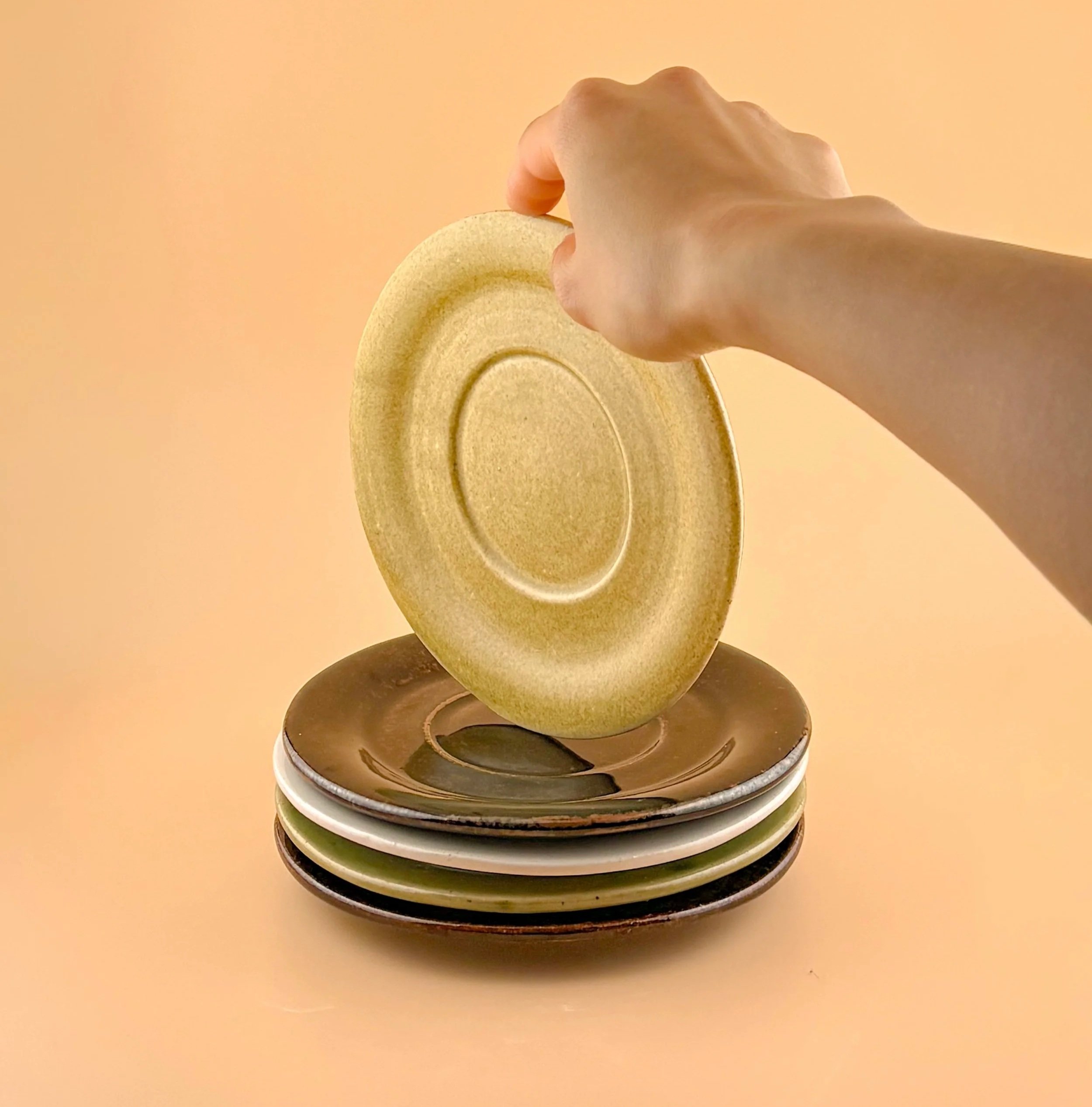 Hand lifting a handmade ceramic saucer from a stacked set, showing scale and glaze texture.