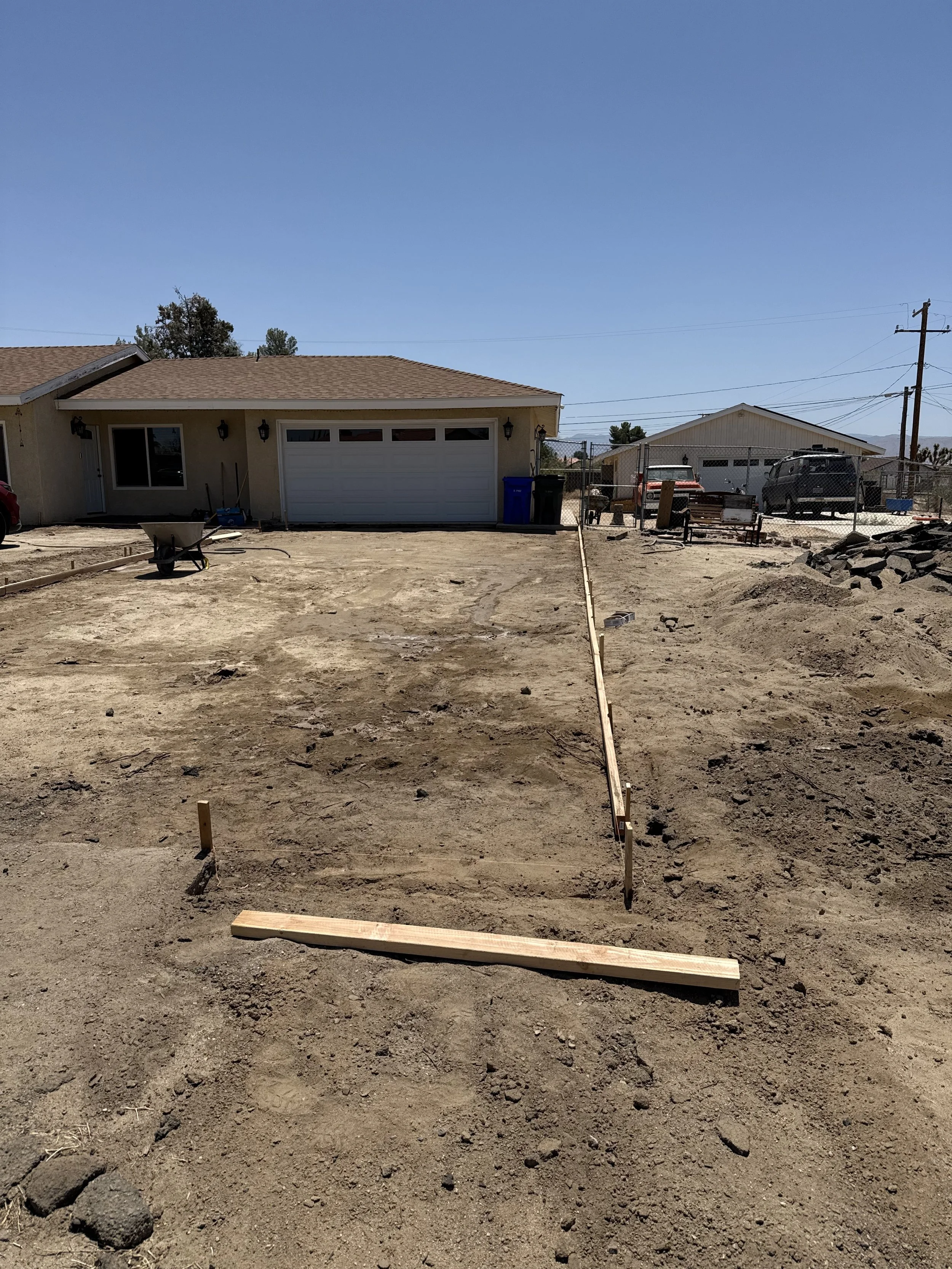 A construction site in front of a house, with framing and ground work in progress for a yard or driveway, includes wooden stakes and a leveling board.
