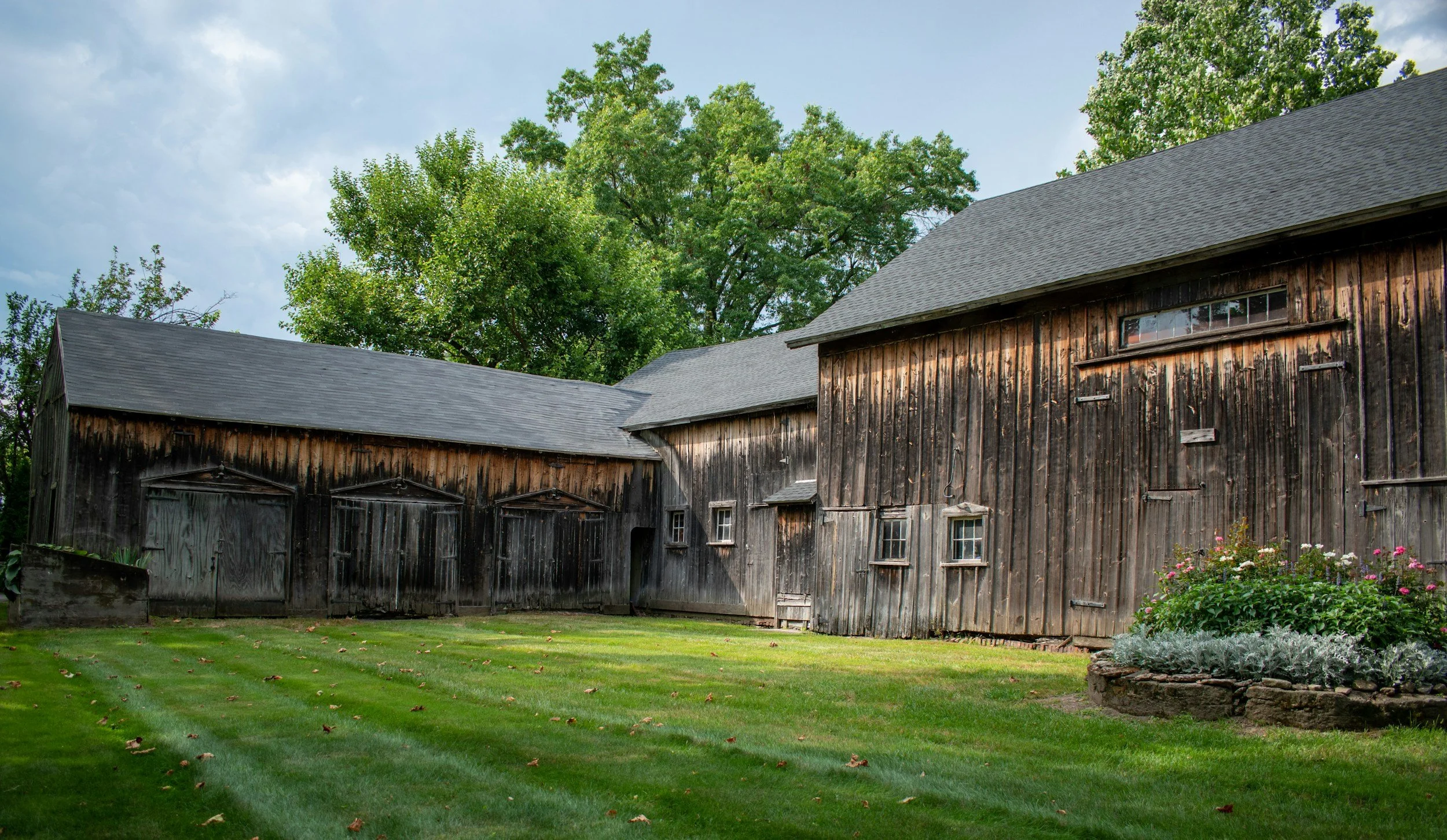 A rustic wooden barn with a gray shingled roof, surrounded by green grass, trees, and a flower bed with pink and white flowers.