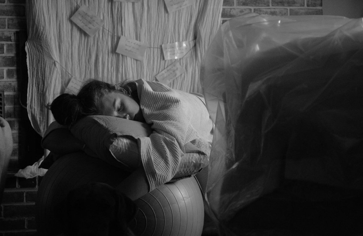A woman sleeping on a large exercise ball, resting her head on a pillow, with notes attached to a fabric-covered surface behind her.