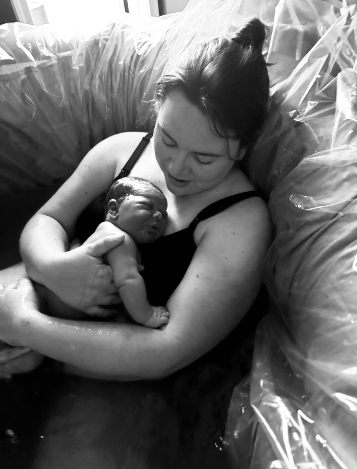 A woman holds a newborn baby in her arms during labor, on a hospital bed with plastic sheeting, smiling and looking down at the baby.