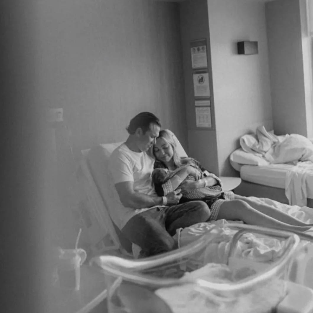 A black and white photo of a couple holding their newborn baby in a hospital room, sitting on a bed with pillows and a hospital bassinet in the foreground.