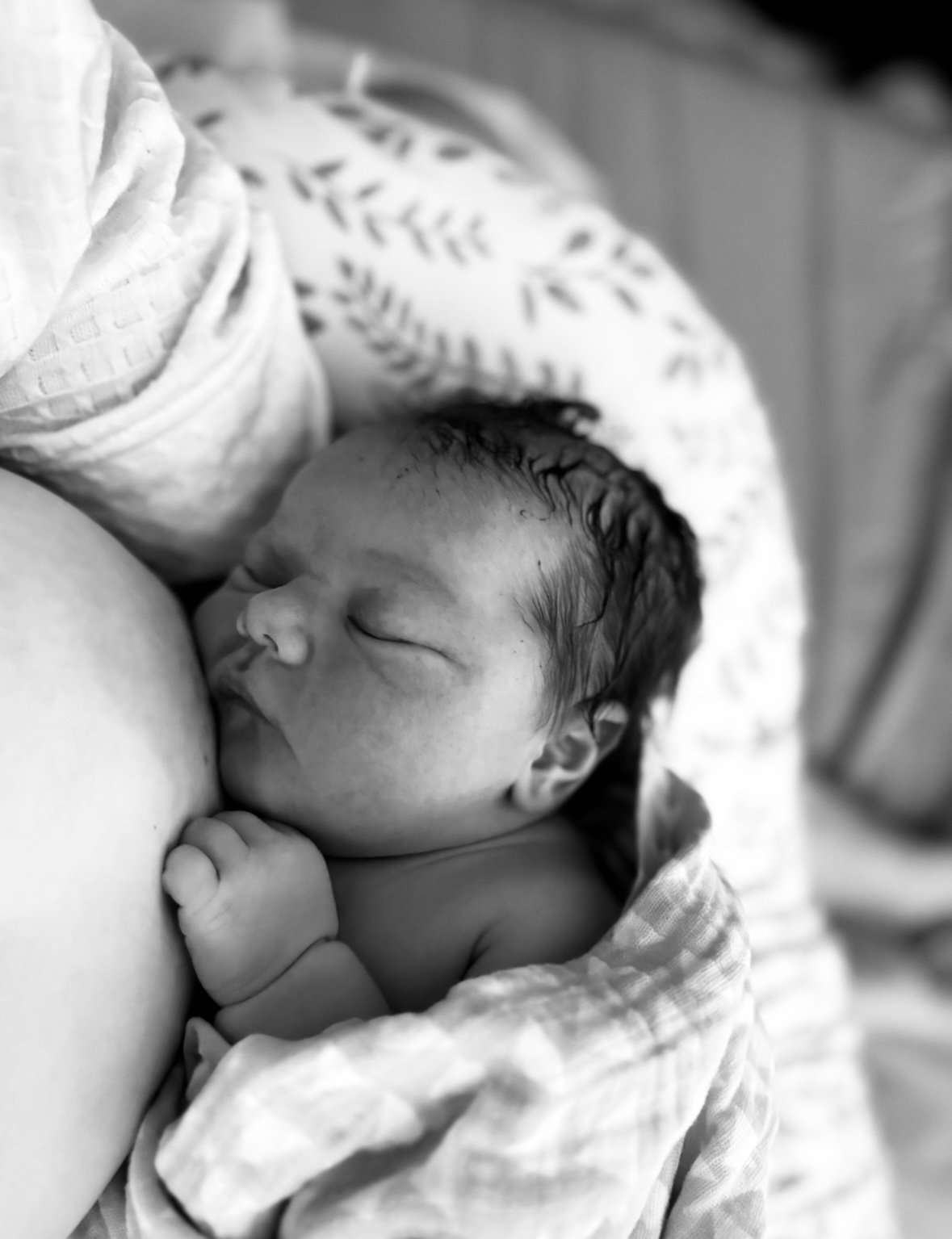 Black and white photo of a newborn baby breastfeeding, with the baby's eyes closed and tiny hand resting near the face.