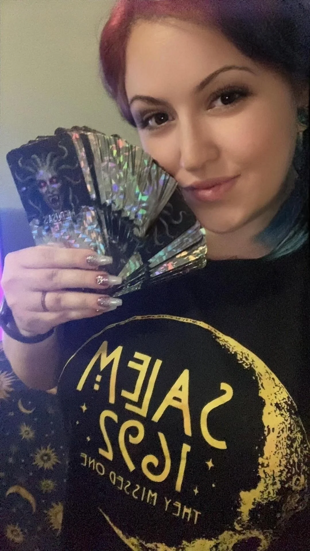 A woman with painted nails holding a fan of holographic tarot cards, smiling slightly, wearing a black shirt with gold moon and star design, and dark hair with purple and teal streaks.