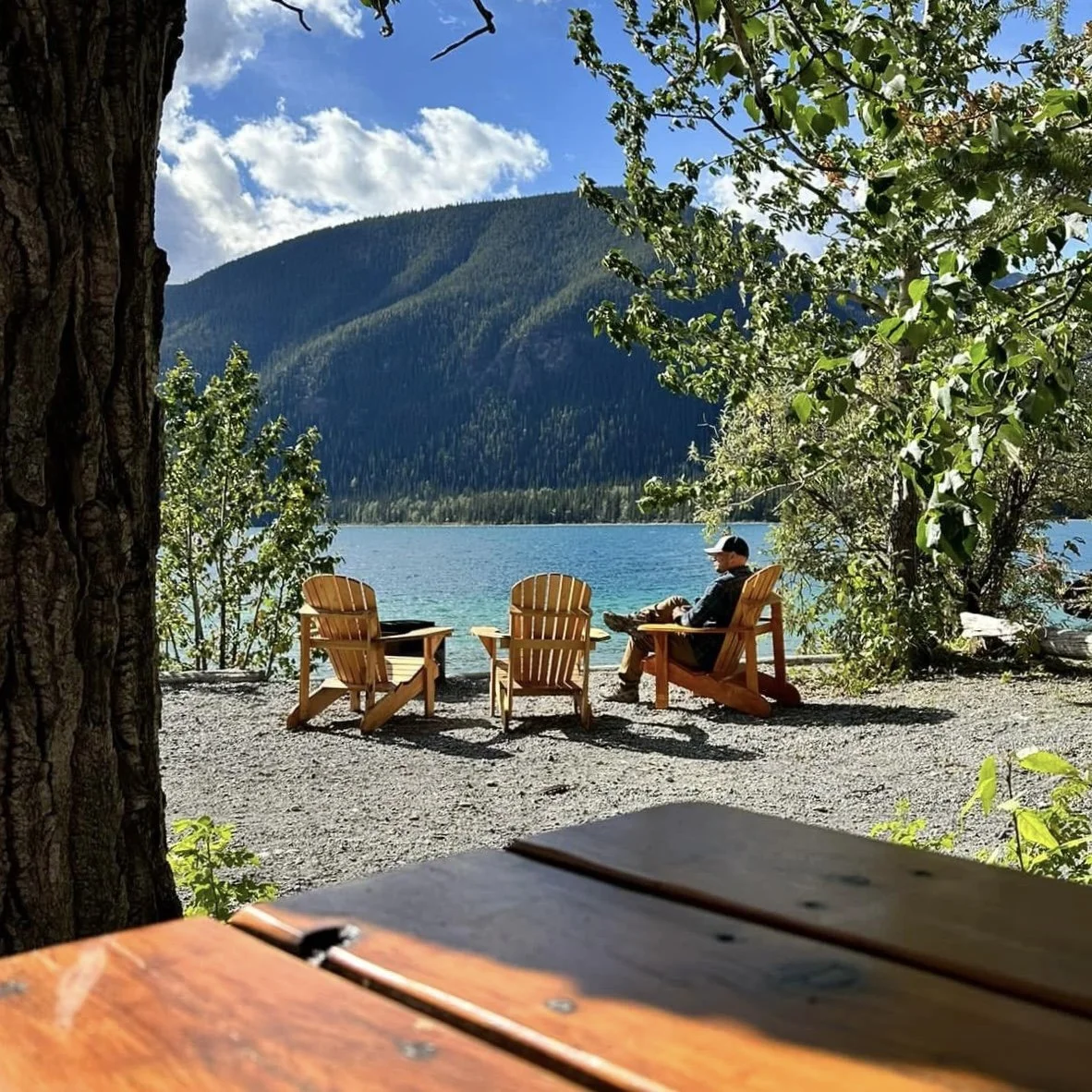 A person sitting in an Adirondack chair by a lake, surrounded by trees and mountains, with three additional chairs nearby, under a partly cloudy sky.
