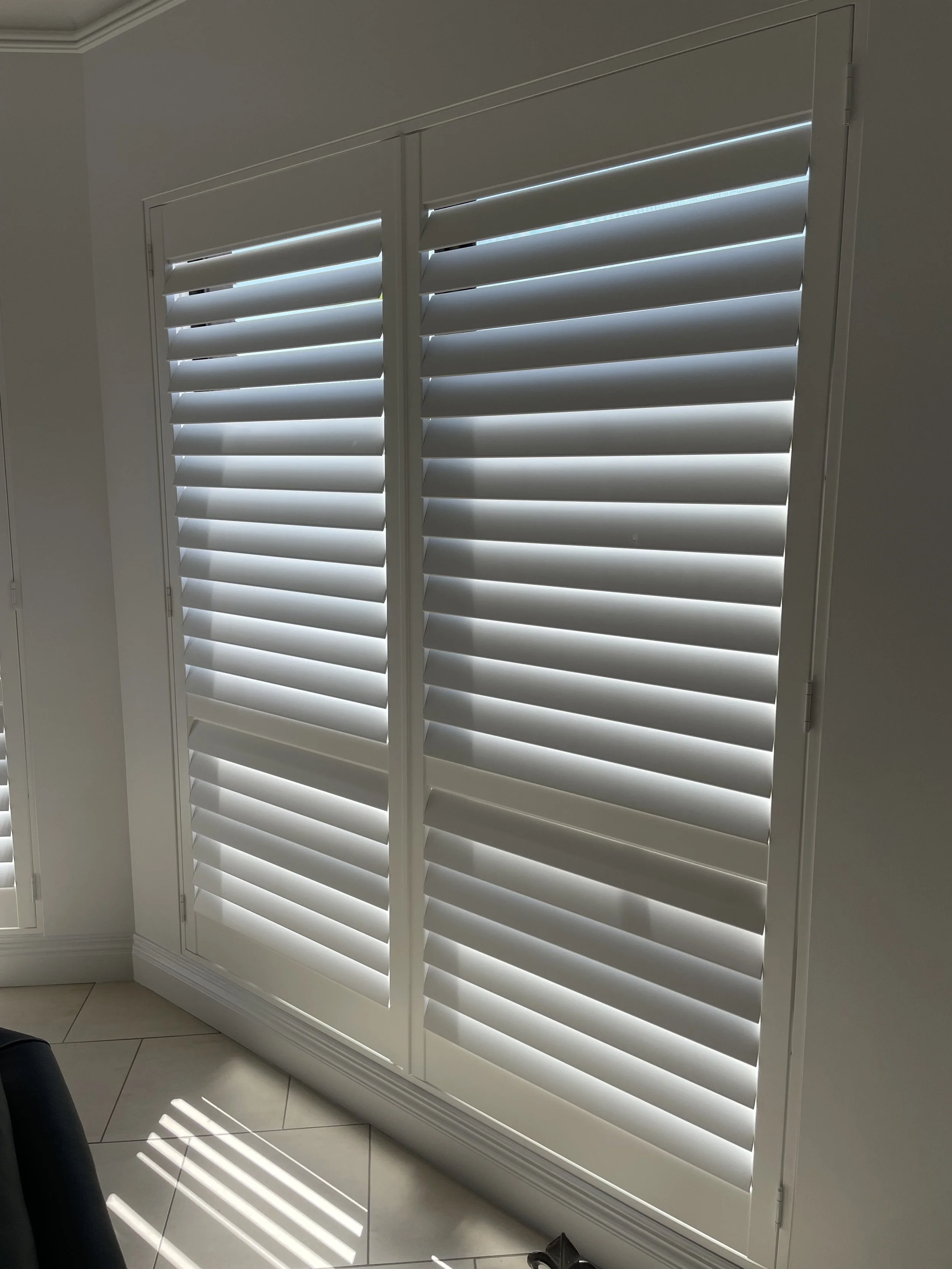 White plantation shutters closed over a glass door, with sunlight casting striped shadows on the tiled floor.