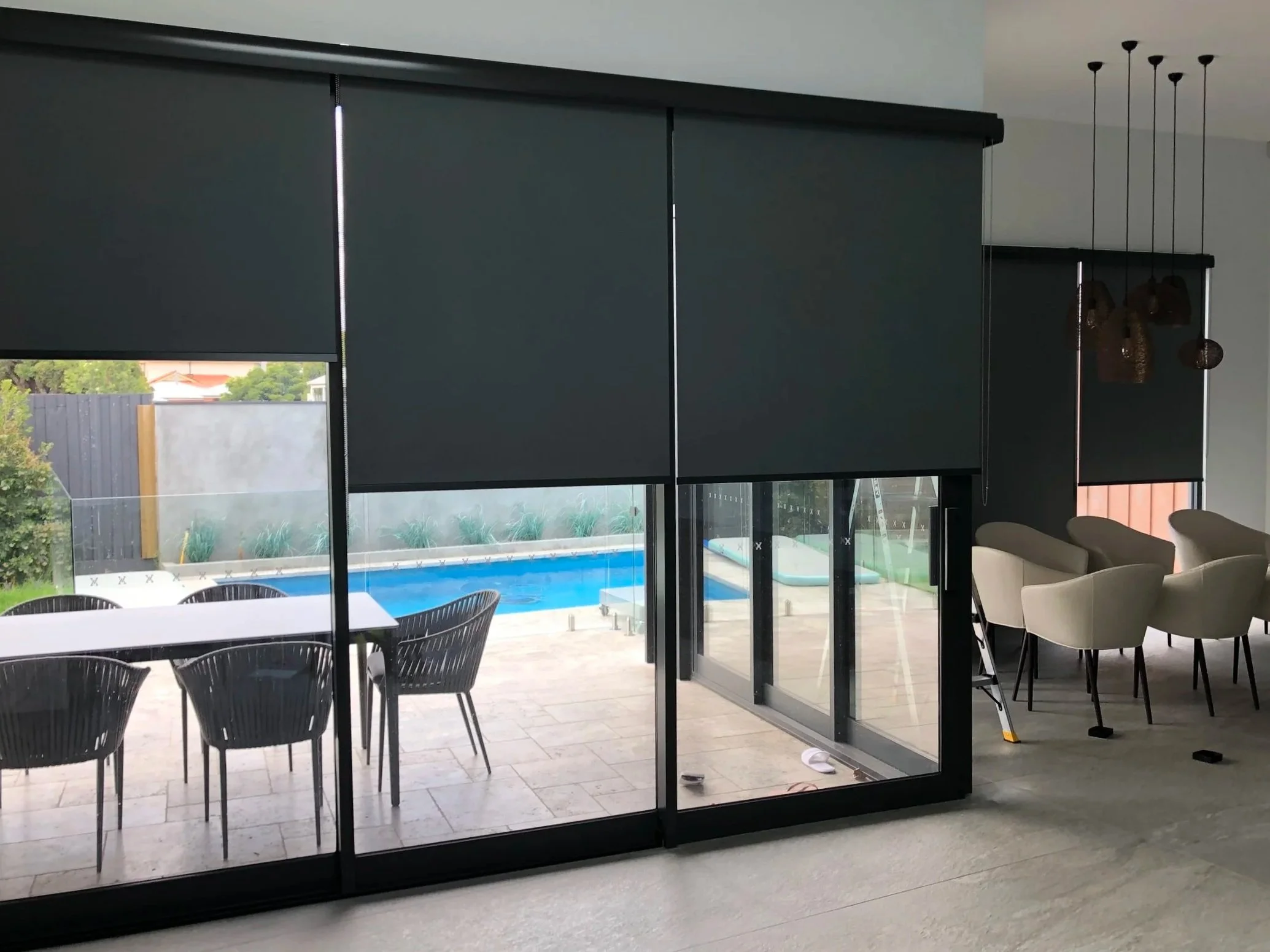 Modern living room with black window blinds, sliding glass door, outdoor patio with dining table and chairs, swimming pool, and interior seating area with white chairs.