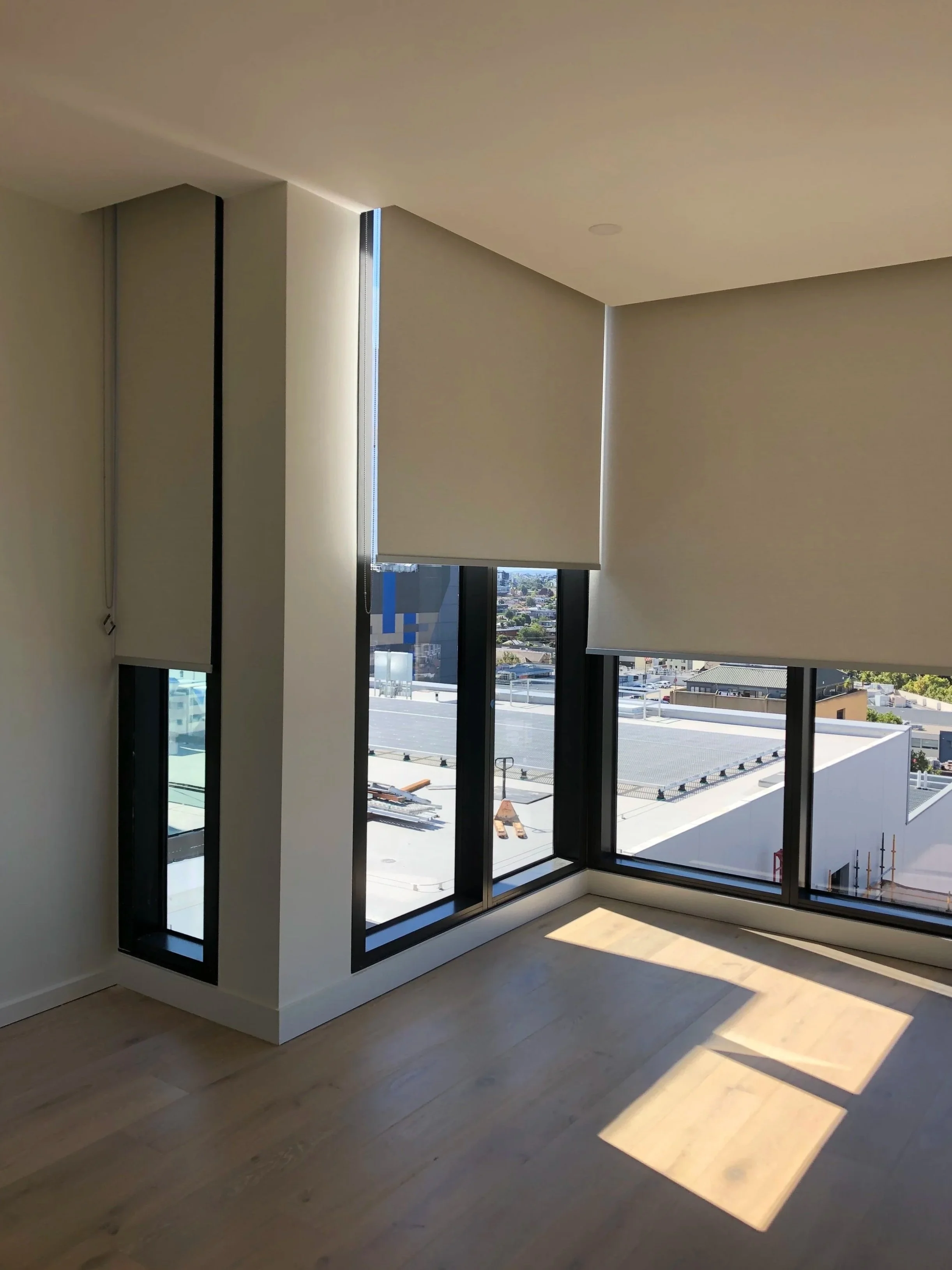 Empty room with large windows and beige blinds, sunlight creates shadow patterns on the wooden floor.