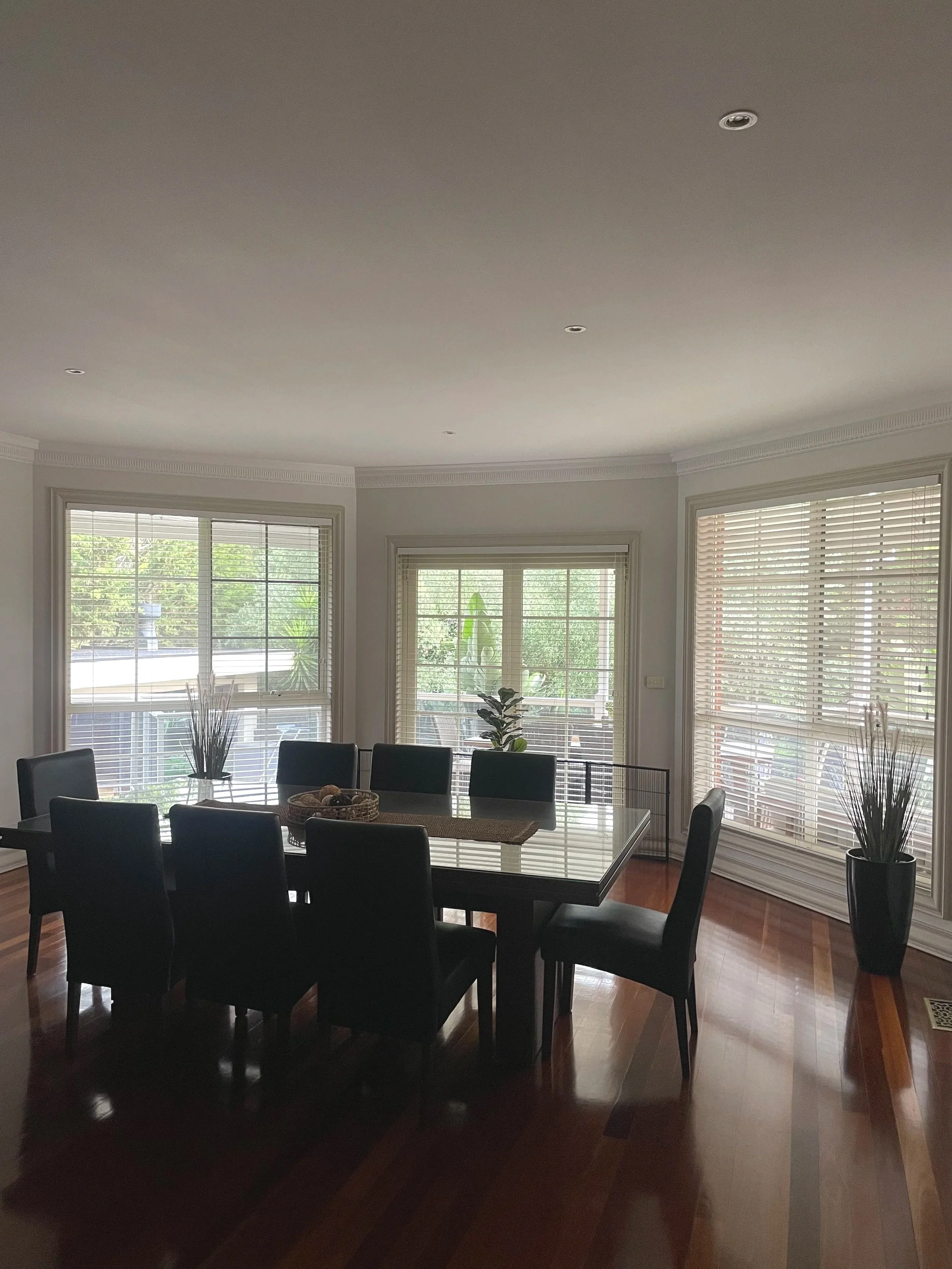 Dining room with wooden floor, black chairs around a glass-top table, large windows with blinds, and potted plants, with sunlight coming in.