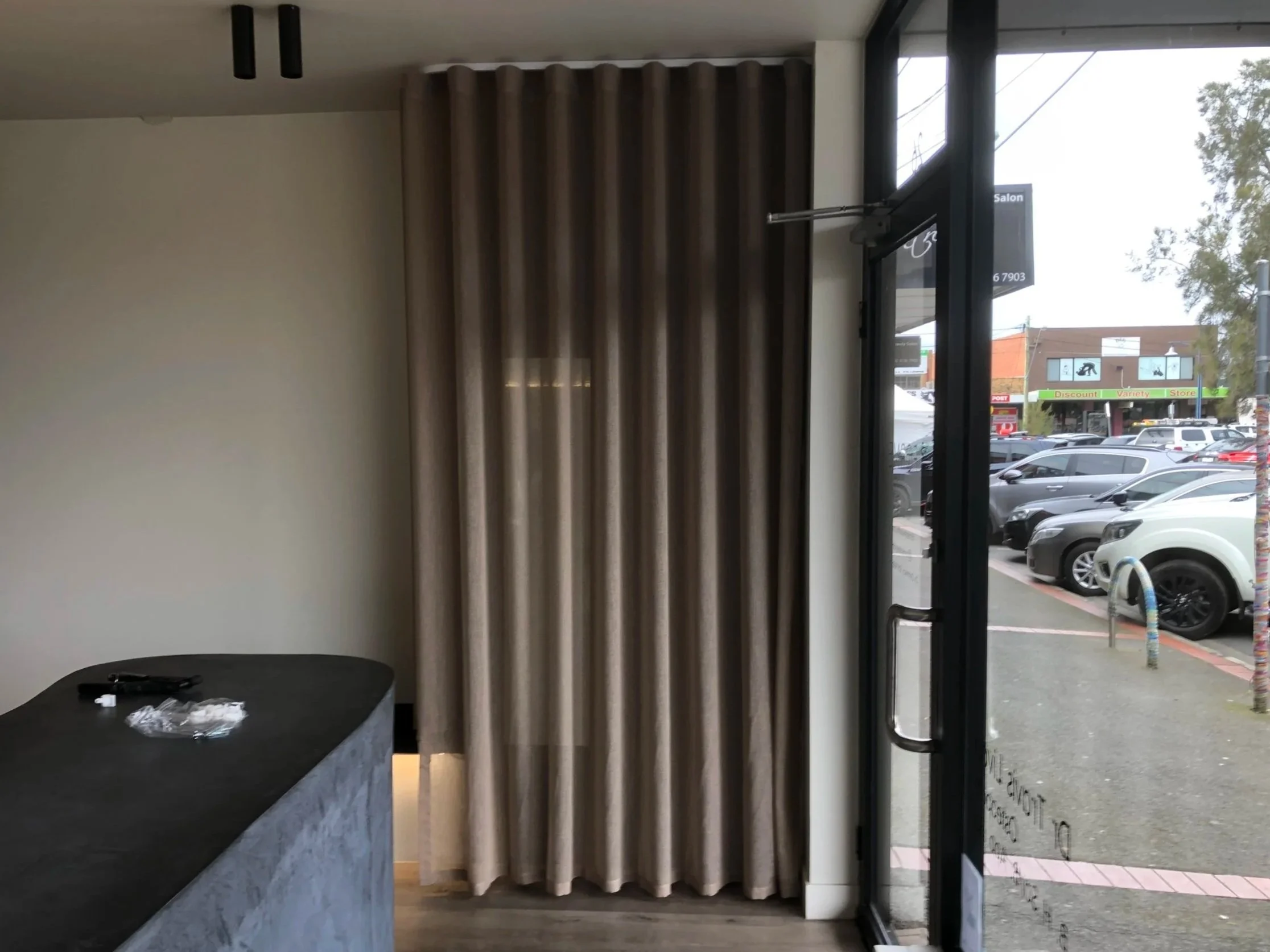 Interior of a store or cafe with a black countertop, beige striped curtains, and glass door leading outside to a parking lot with cars and a strip mall.