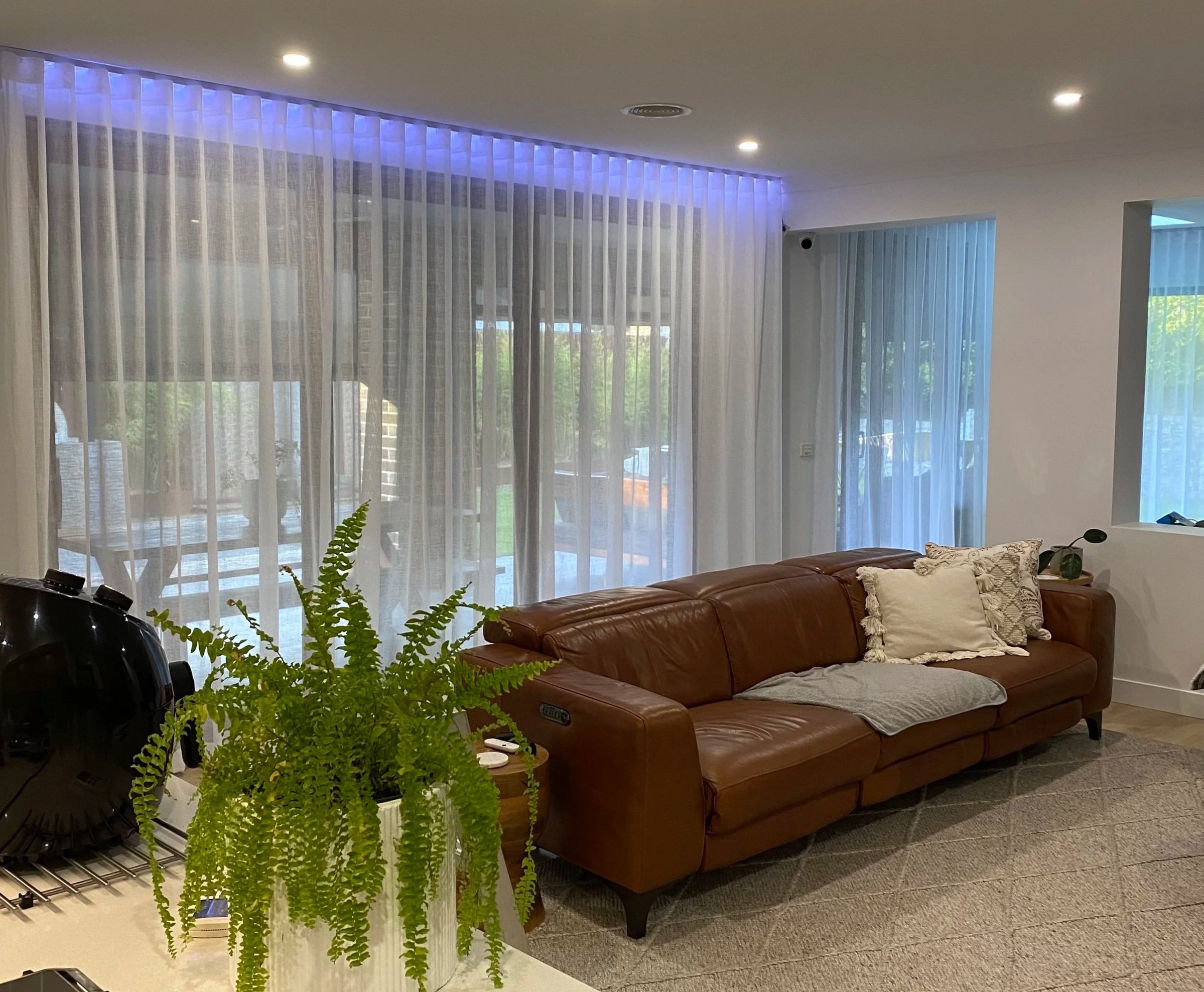 Living room with a brown leather couch, decorative pillows, a potted fern on the table, and sheer curtains covering large windows with outdoor patio visible.