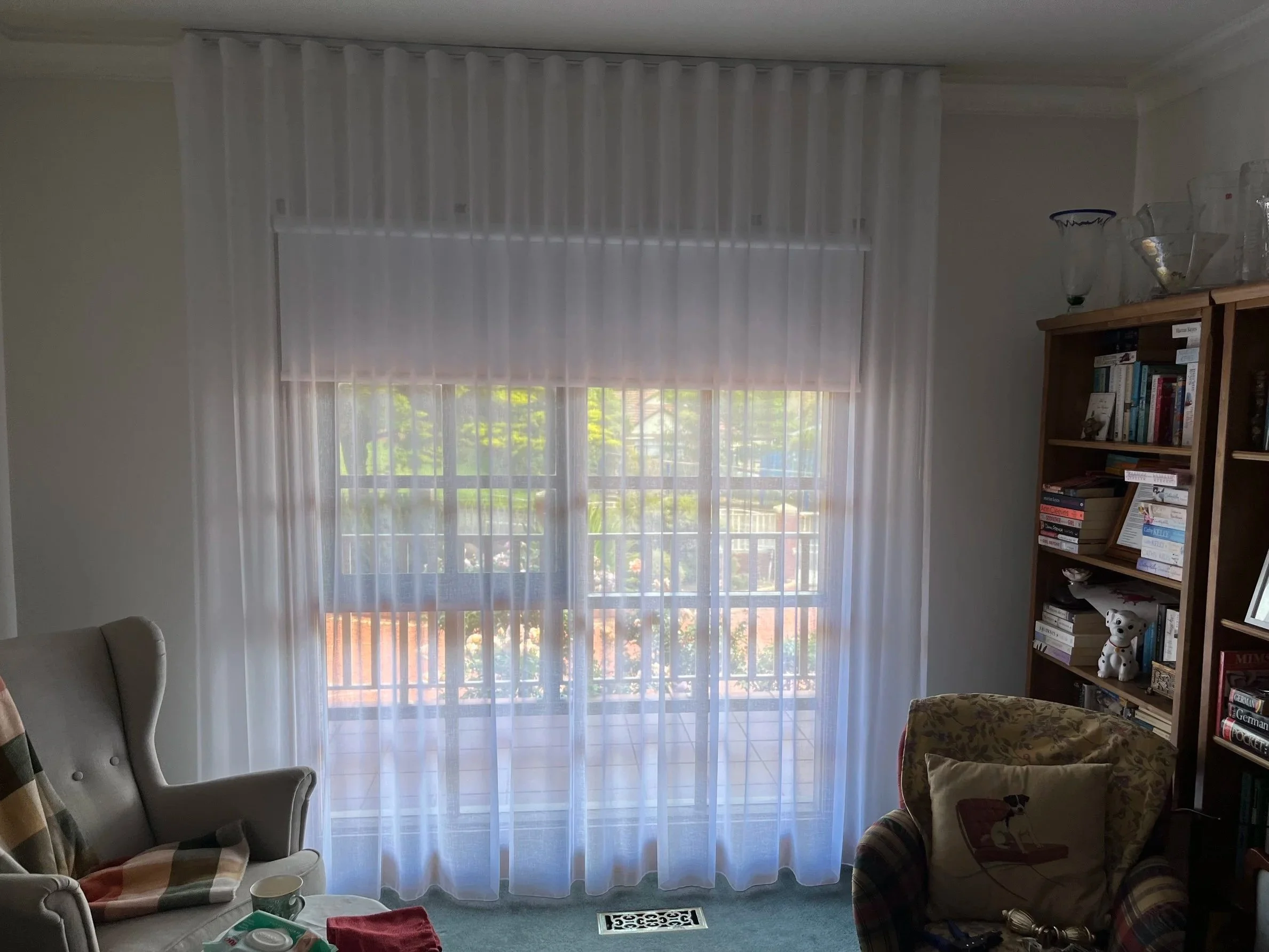 Living room with sheer white curtains covering a sliding glass door, a beige armchair with patterned cushions, a wooden bookshelf filled with books and decorative items, and a small table with miscellaneous objects.