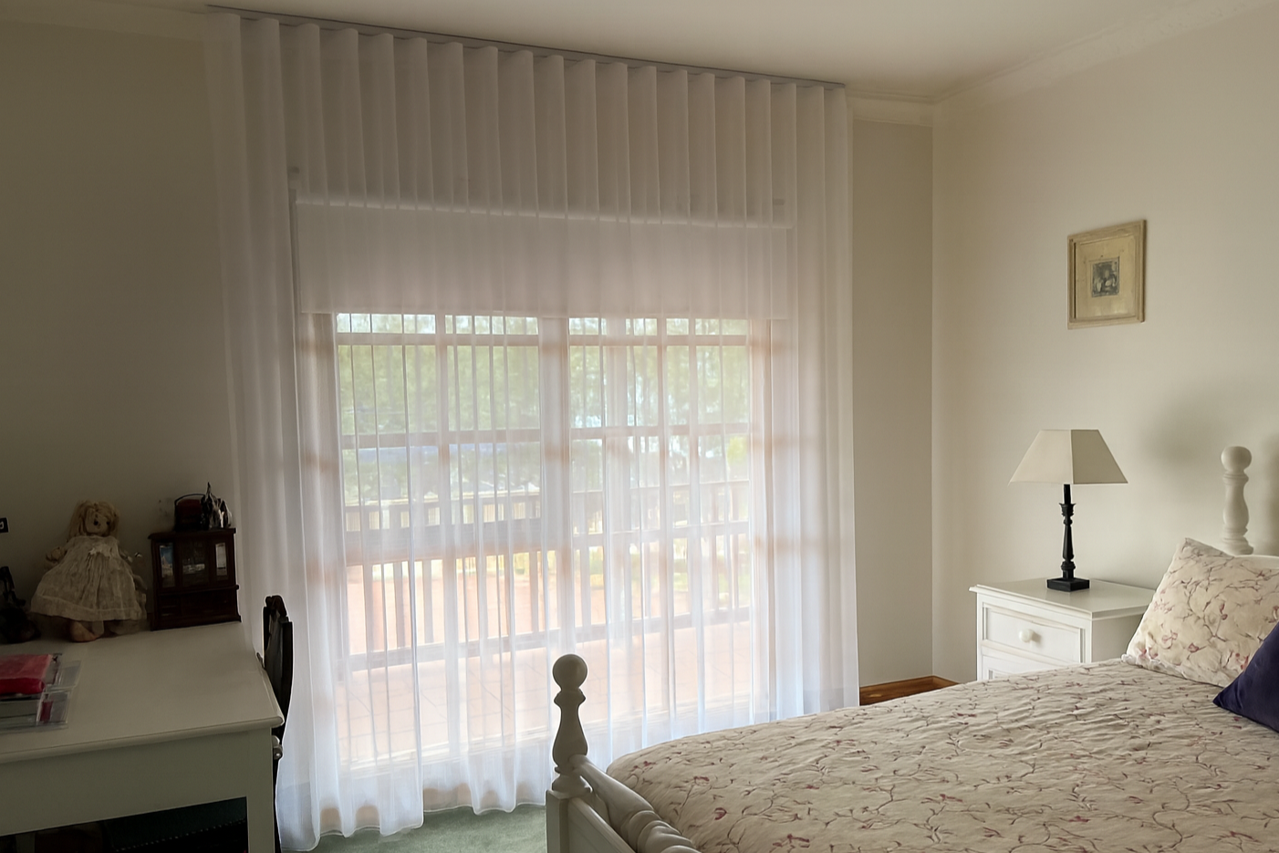 A bedroom with a bed, nightstand, table lamp, sheer curtains, and a window with a balcony outside.