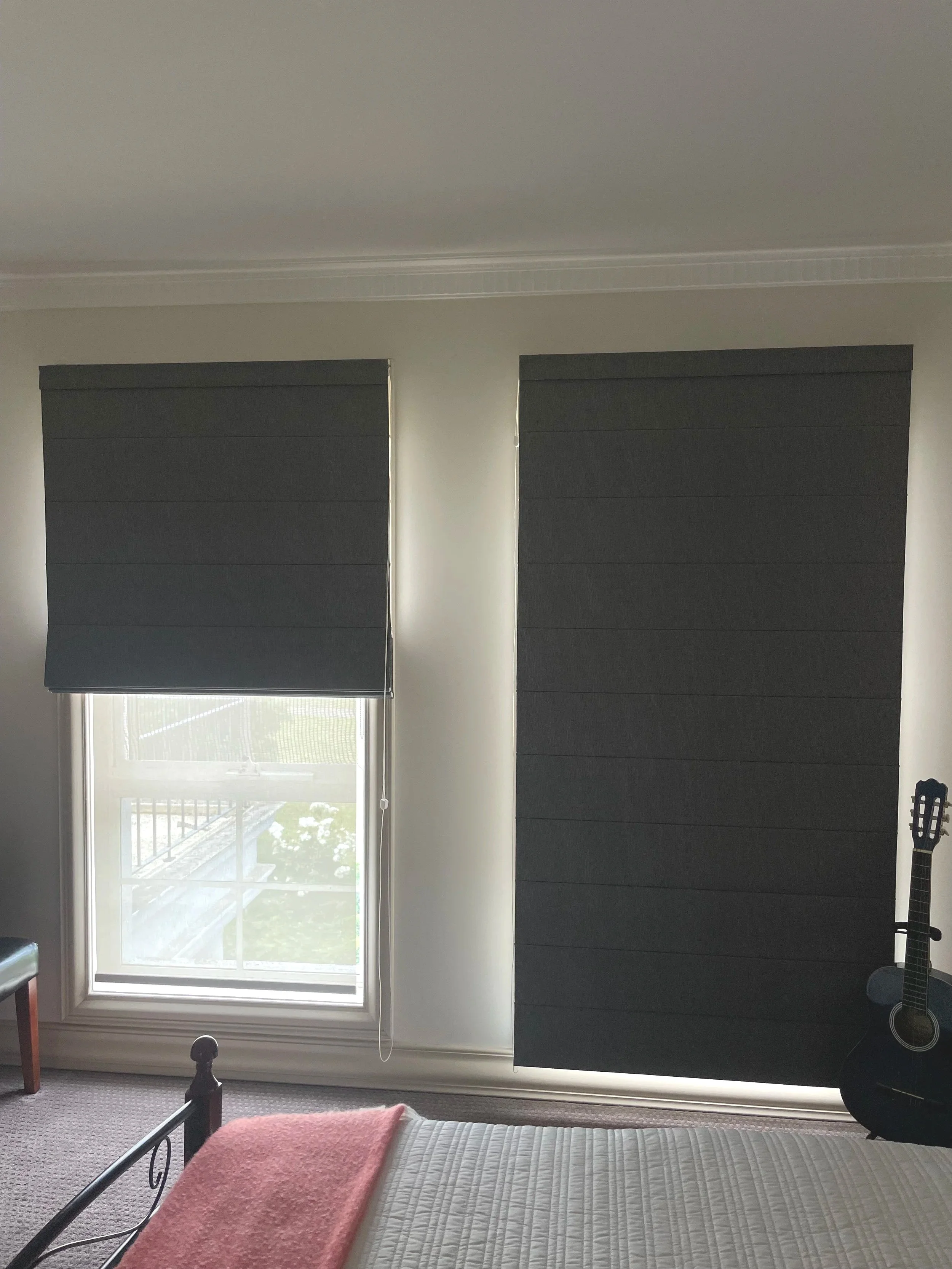 Bedroom with two windows covered by dark grey blinds, a bed with a pink blanket, a guitar on a stand near the right wall, and a black bench near the left window.
