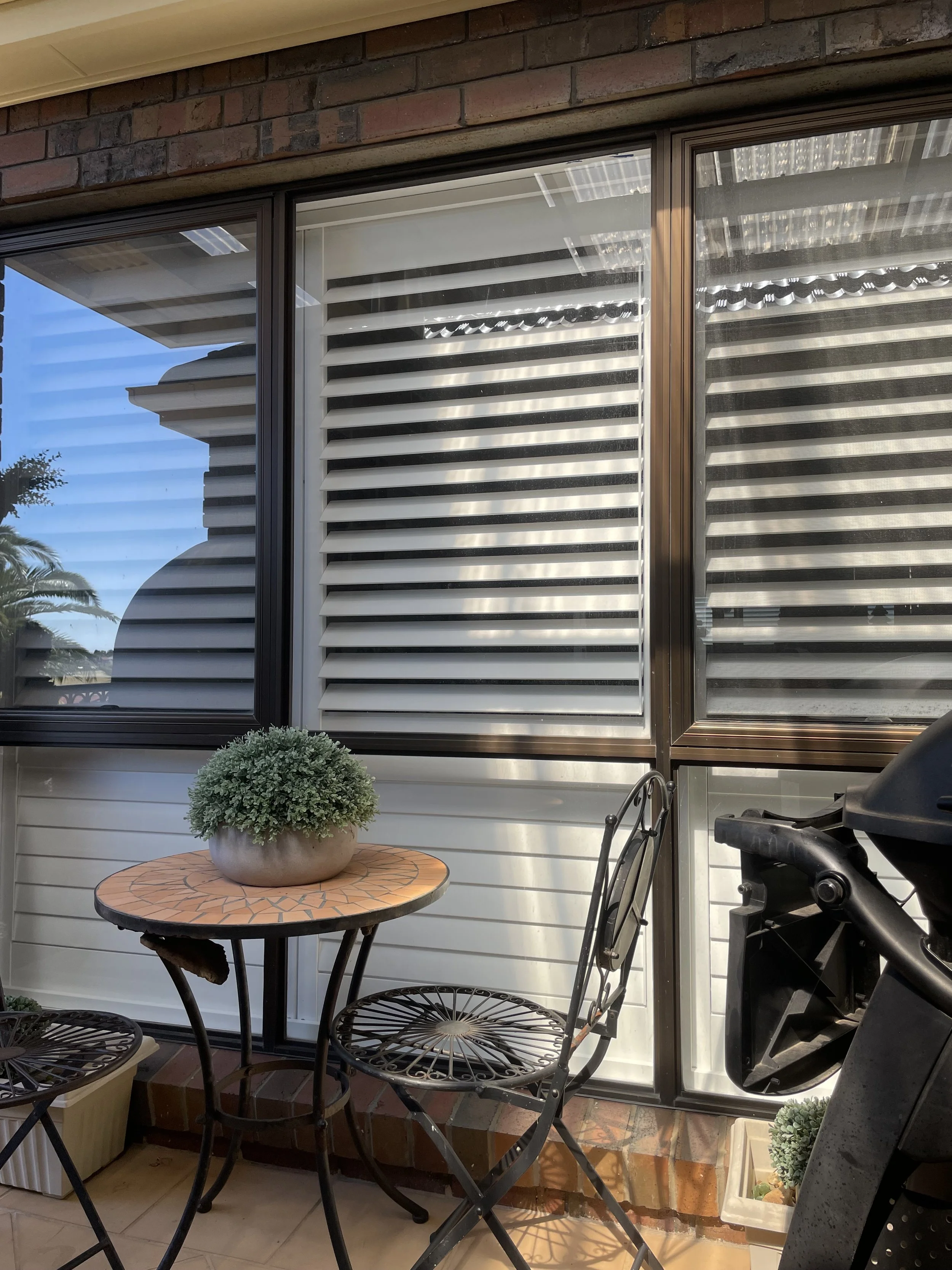 Patio with small round table, metal chairs, potted plant, and grill in front of a window with adjustable shutters, brick wall, and outdoor view.