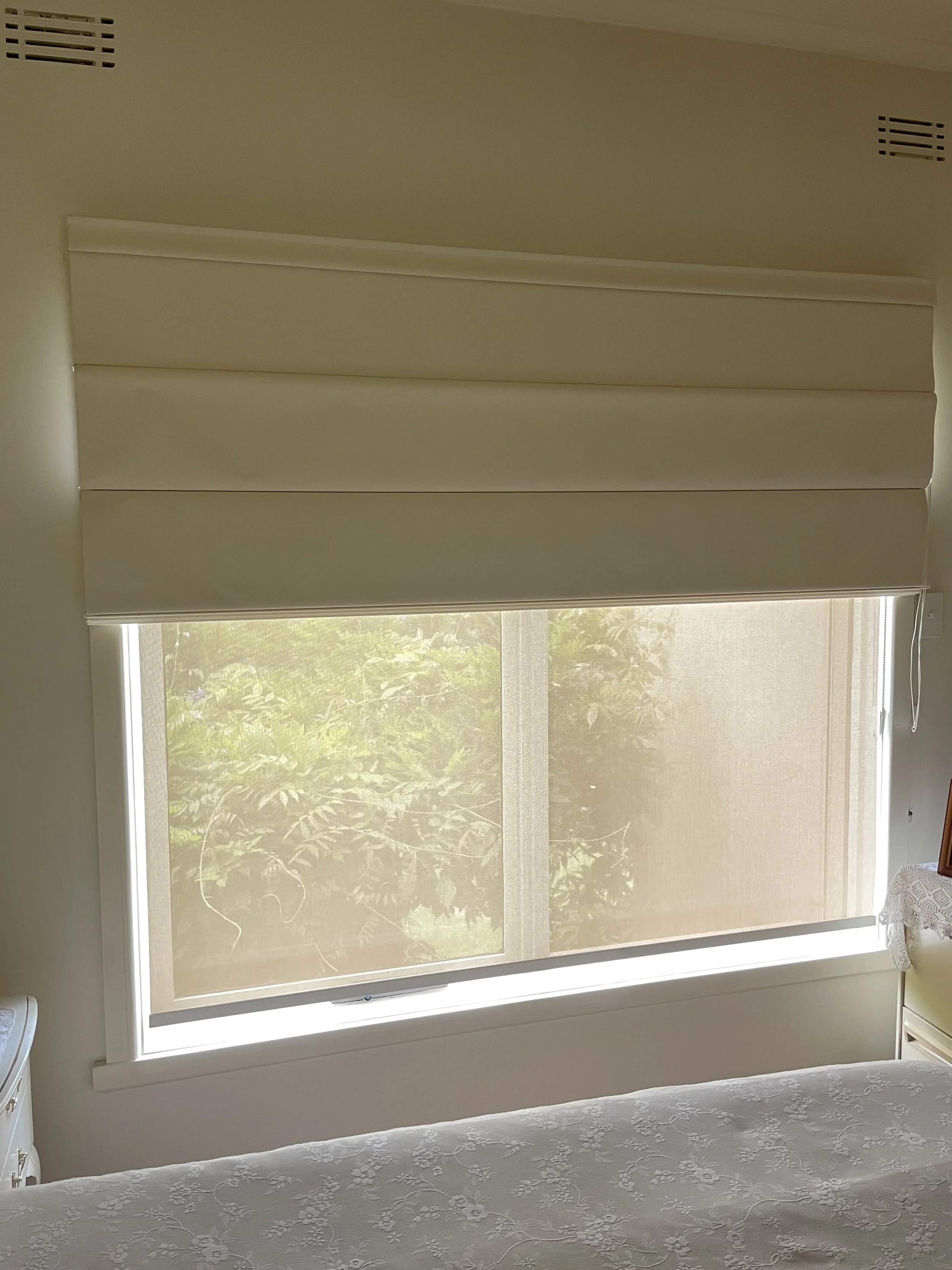 A window with beige blinds partially pulled down, showing green foliage outside.