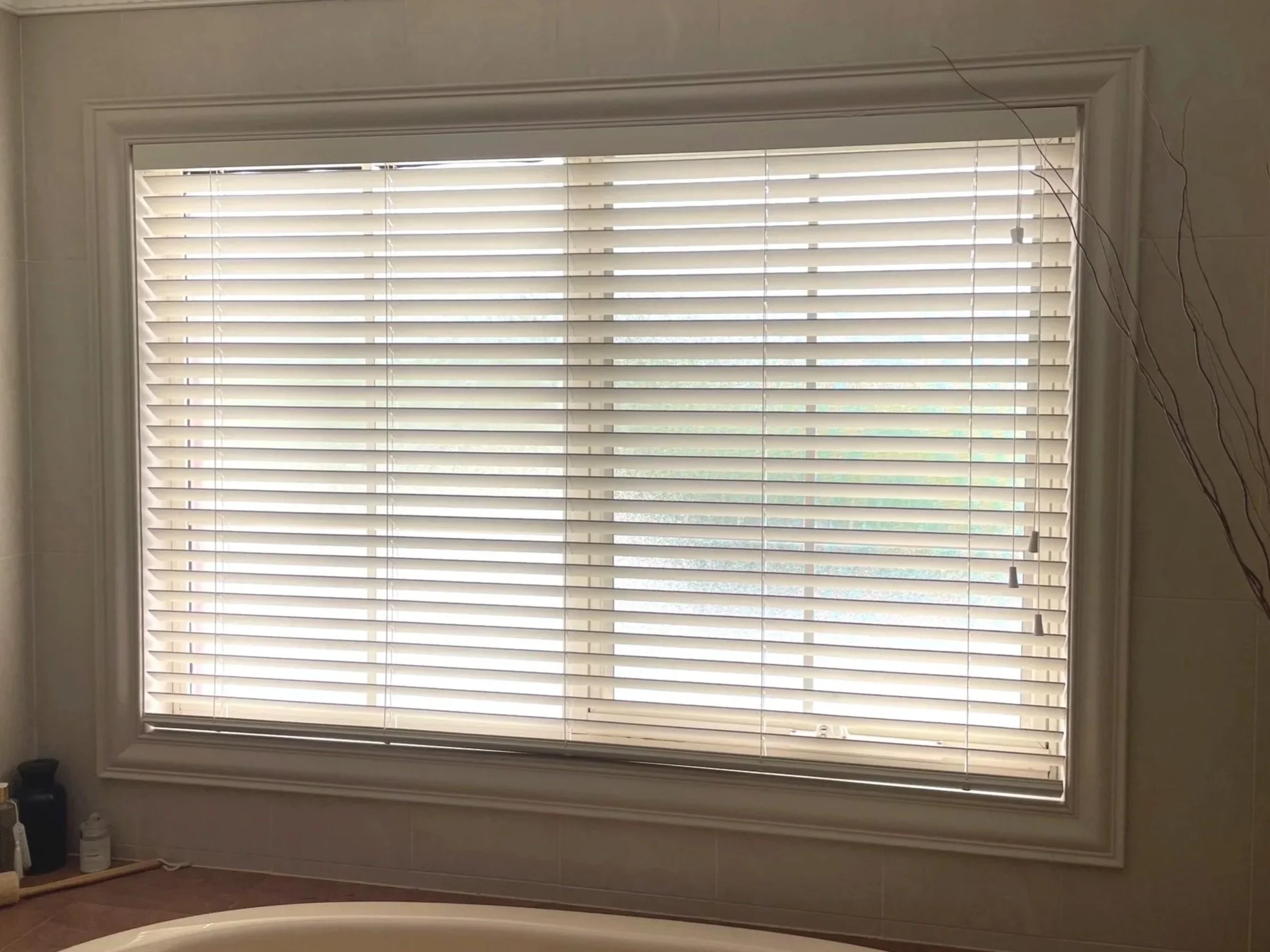A large window with white horizontal blinds, situated above a bathroom bathtub. The window is framed with decorative molding, and some thin, wavy twigs are on the right side of the window.