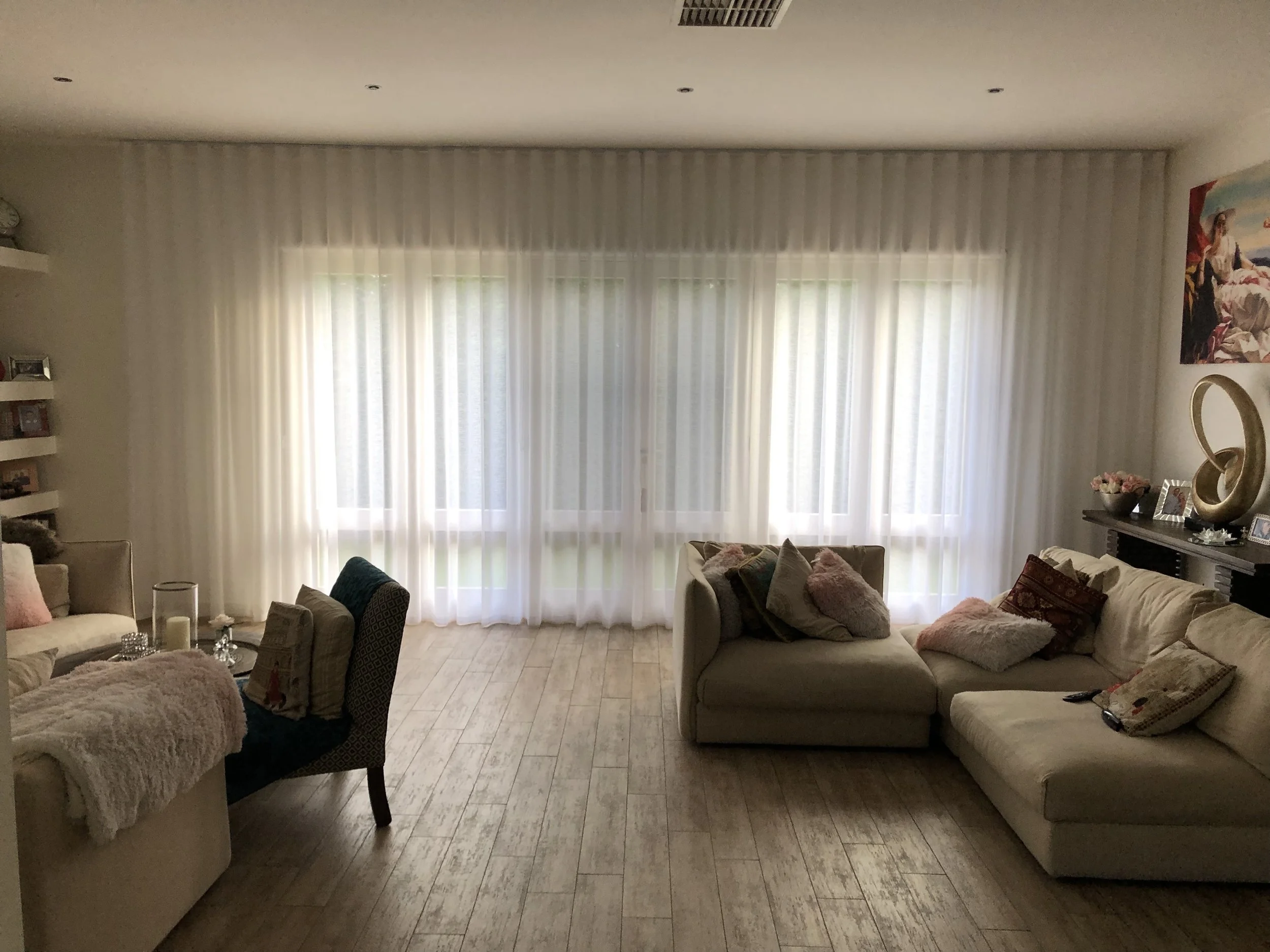 Living room with cream-colored sofas and armchair, decorated with pillows, beige curtains, and a wooden floor. There are shelves with pictures on the left, a fireplace with decorative items, and artworks on the wall.