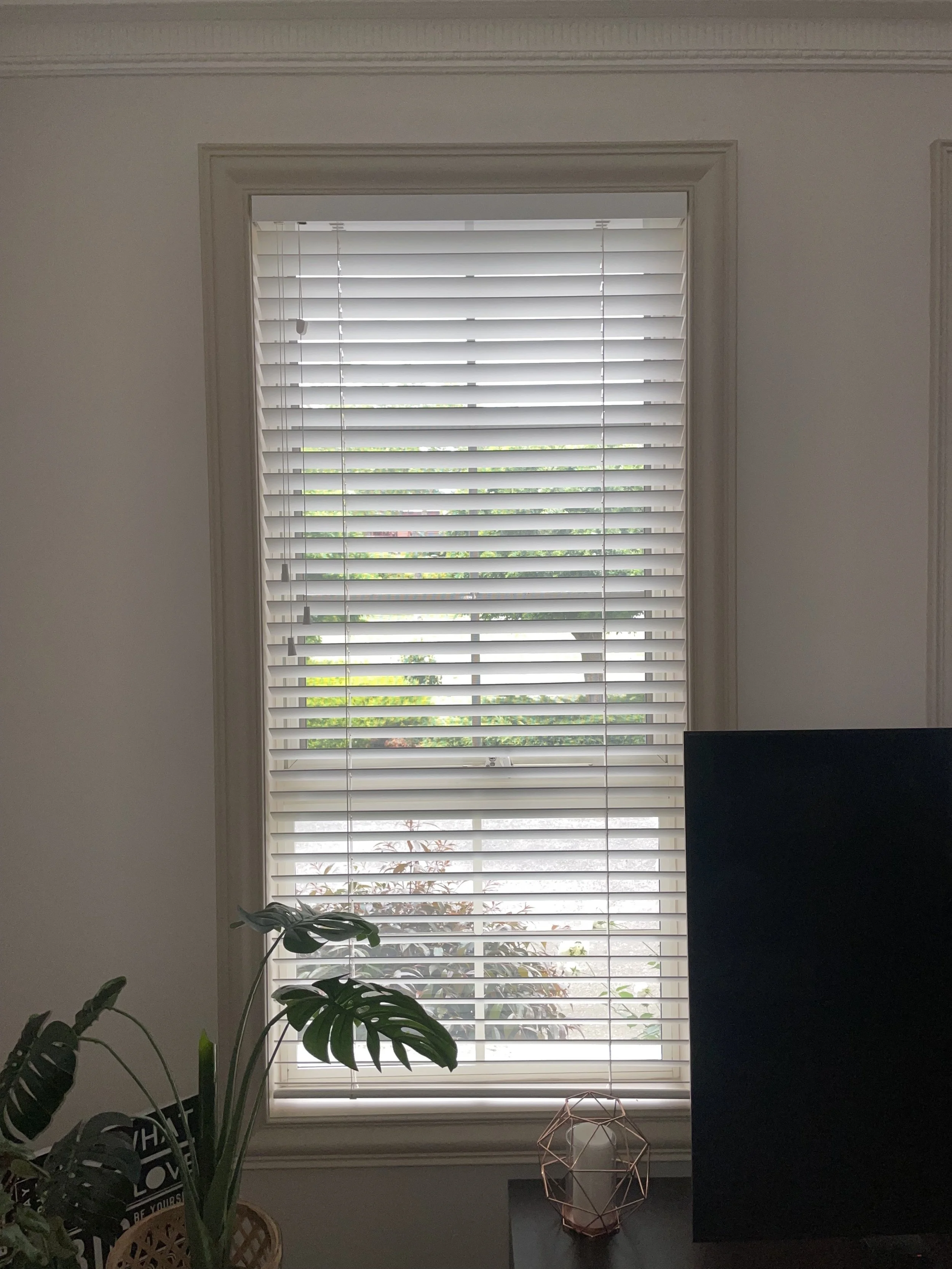 A window with white blinds partially open, revealing greenery outside. The window frame is beige, and the wall surrounding it is white. In front of the window, a television screen is visible on the right side, and a table with a decorative geometric 