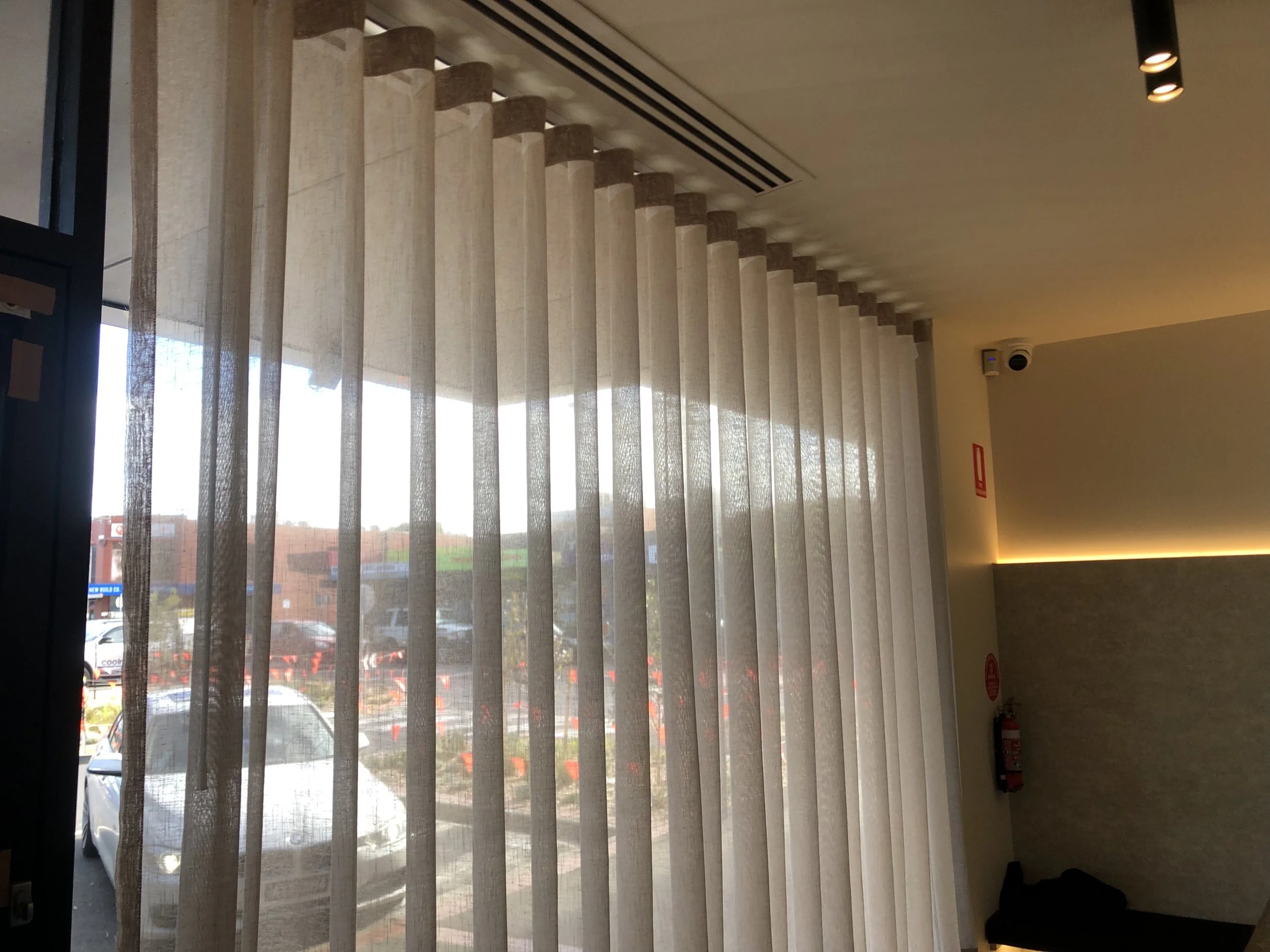 Interior of a room with large window covered by sheer beige curtains, a fire extinguisher on the wall, and a partially visible black object near the corner.