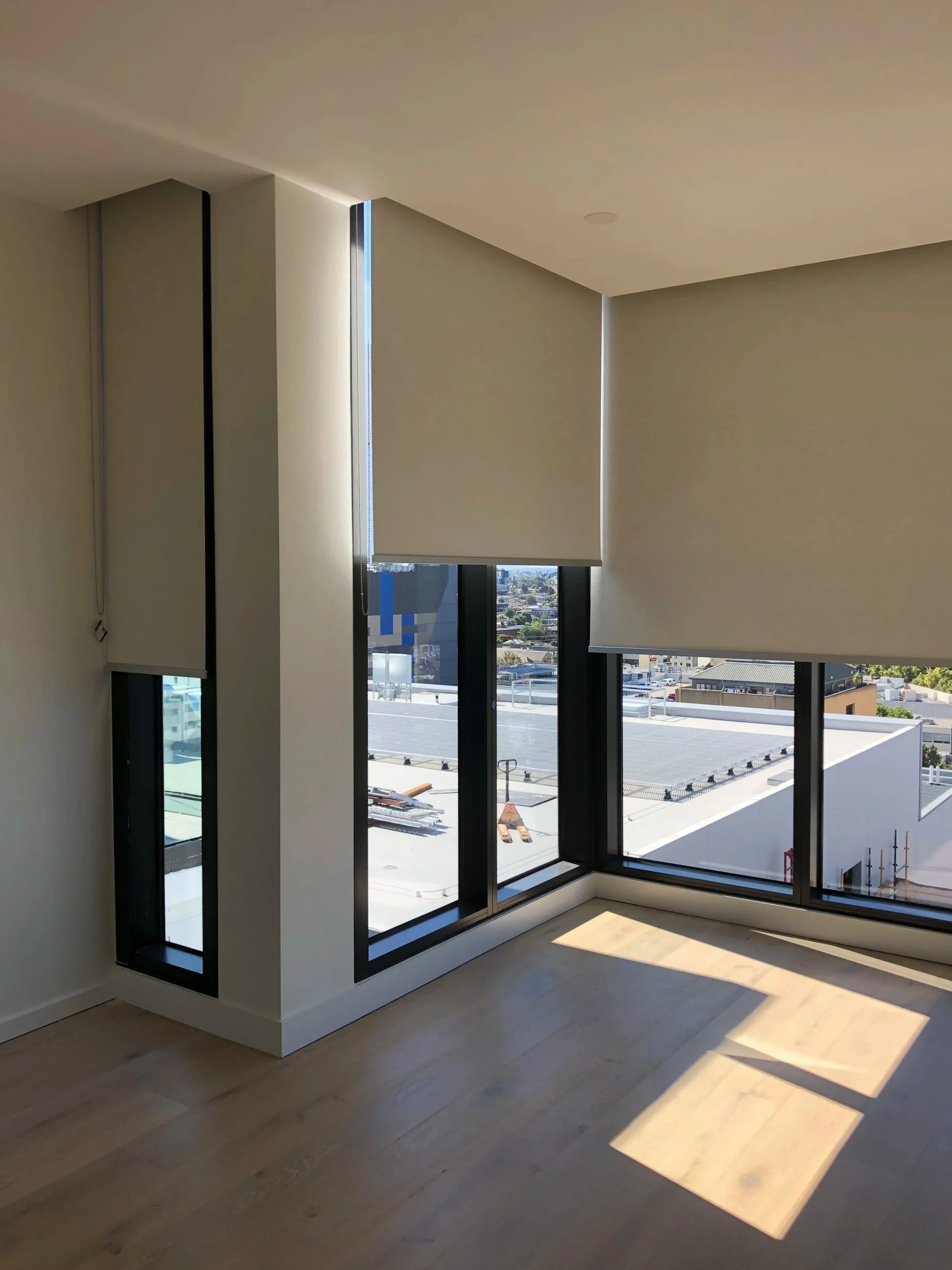 Empty room with large windows and roller blinds, sunlight casting shadows on the wooden floor.