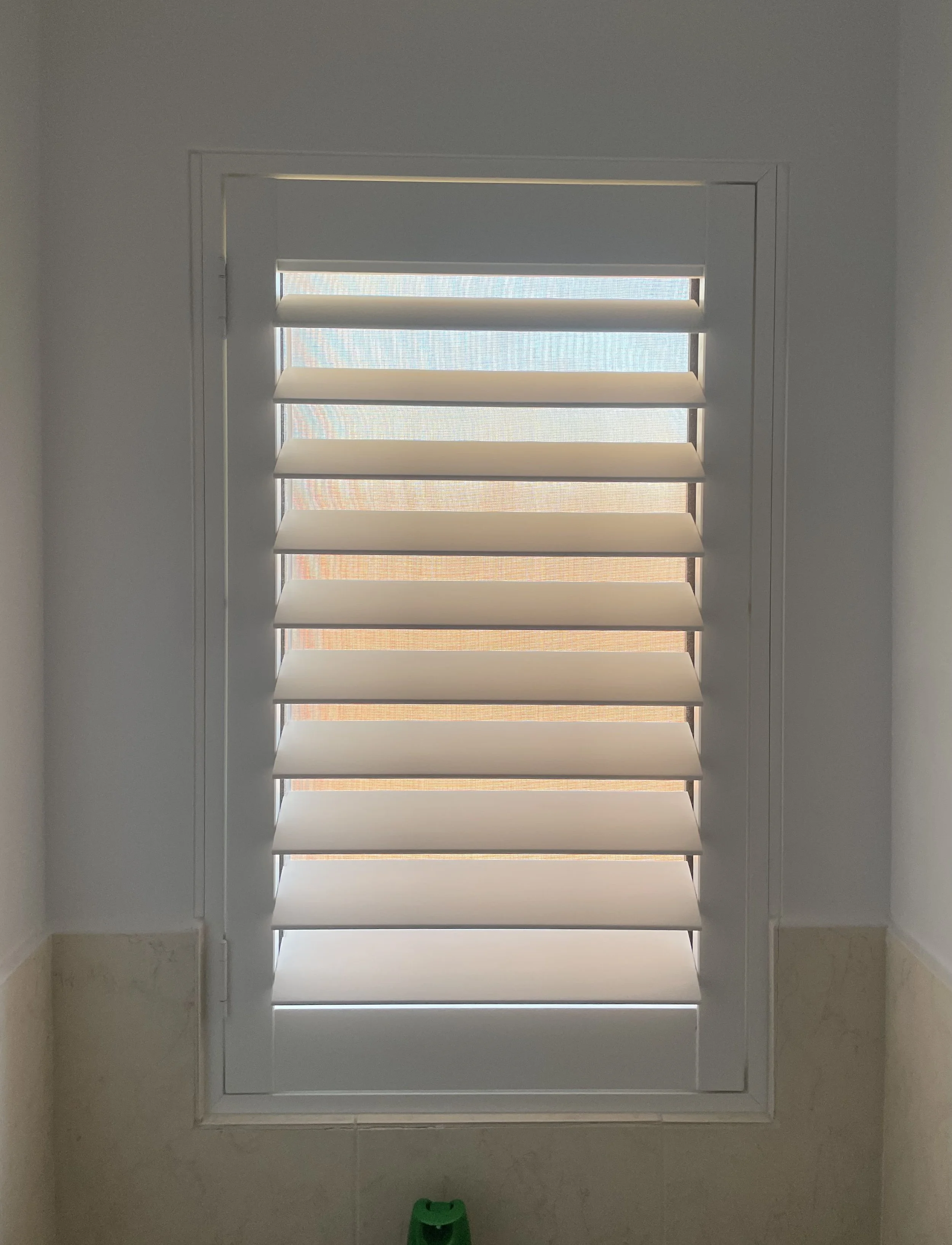 A window with white plantation shutters in a room with white walls and beige tiled lower walls.