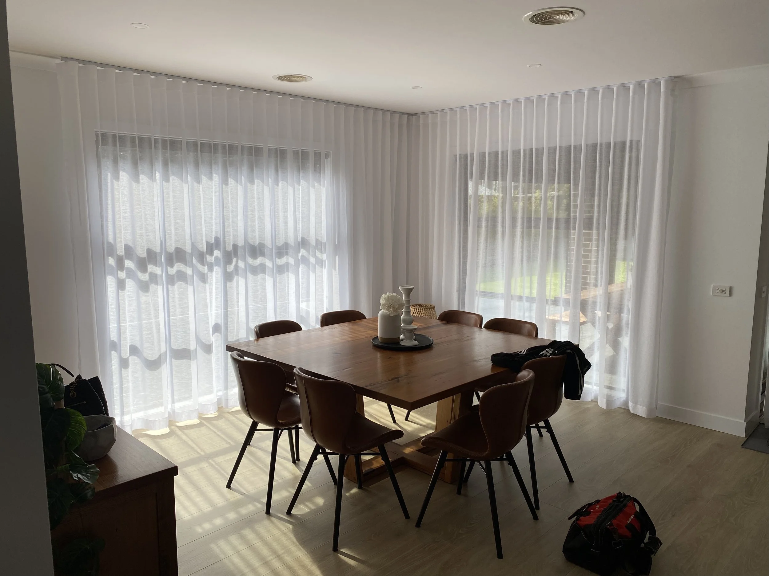 Bright dining room with a wooden table and eight chairs, white sheer curtains on large windows, decorated with candles and flowers, with a black backpack on the floor and a small wooden side table with a plant.