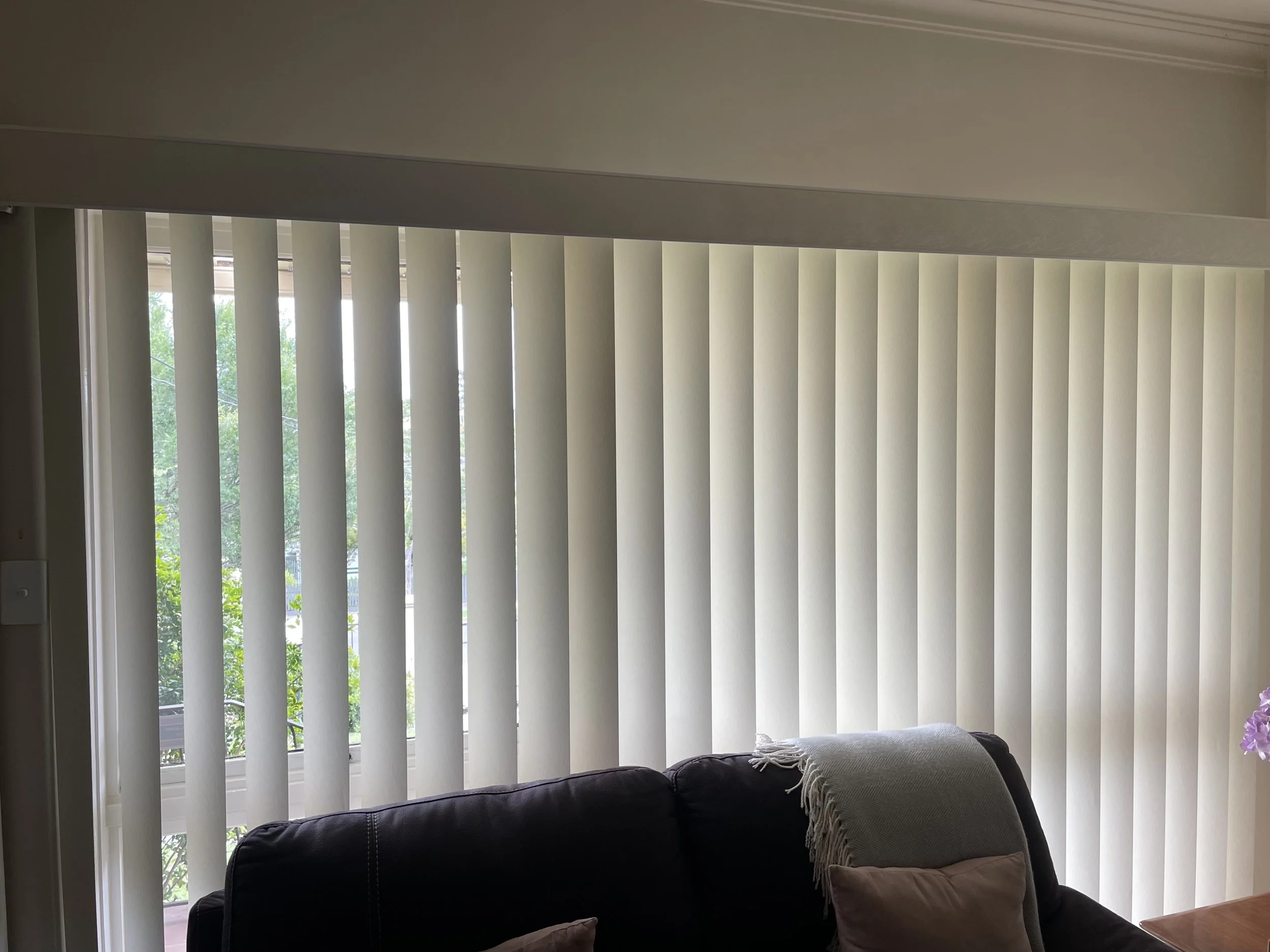 A living room with vertical beige blinds covering a large window, a dark-colored sofa with beige pillows and a beige throw blanket.