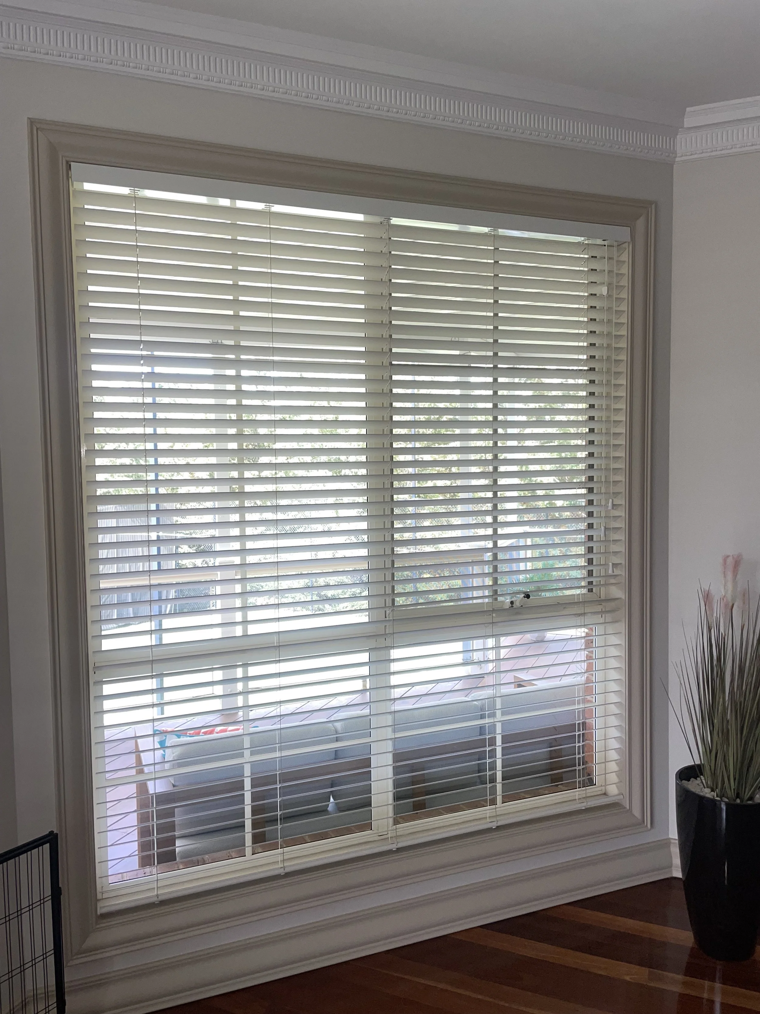 Close-up of a large window with white horizontal blinds, framed with beige molding, overlooking a balcony with outdoor furniture, indoors on a hardwood floor, with a tall black planter containing ornamental grass on the right.