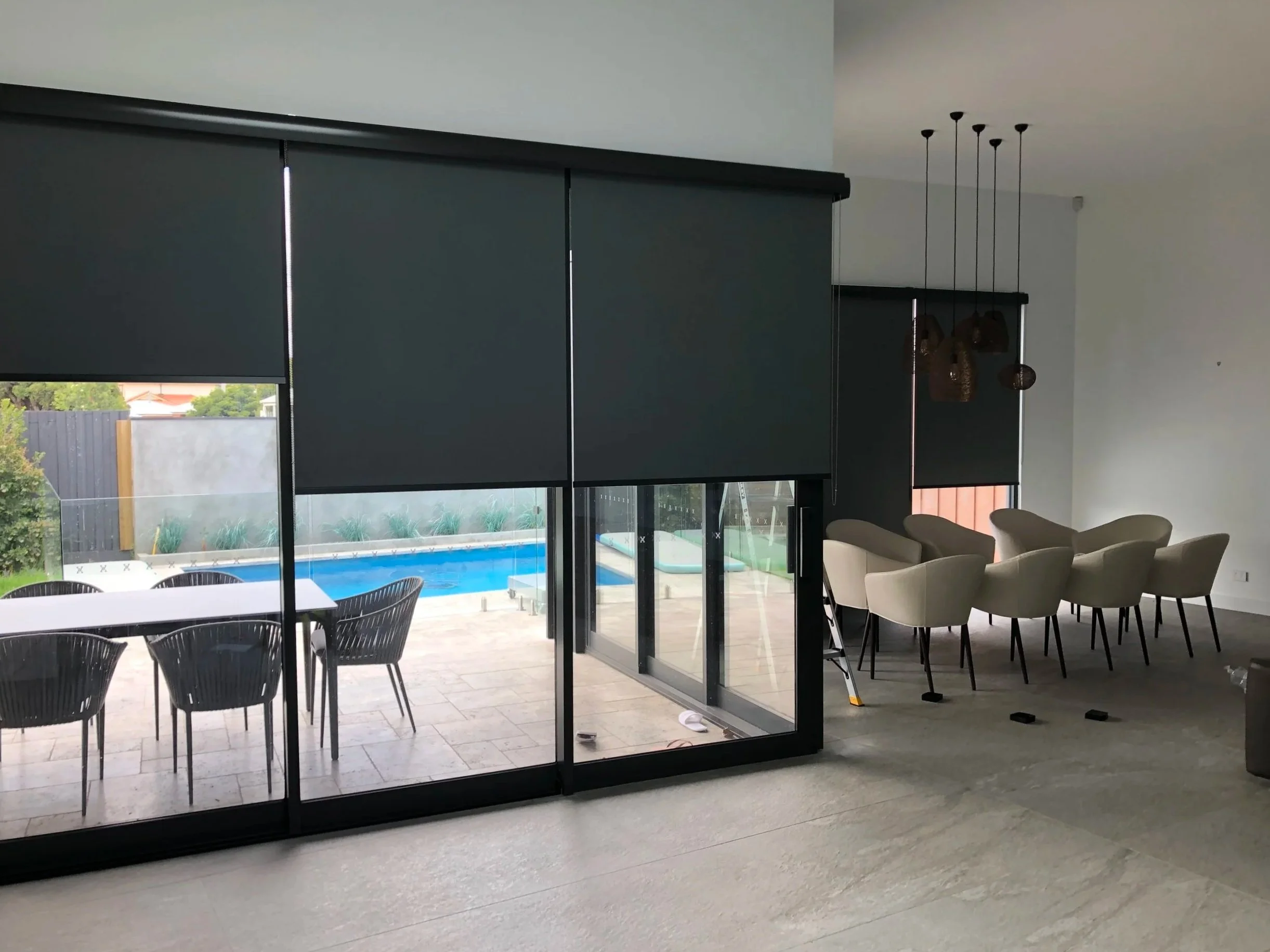 Interior of a modern living space with black window blinds, a view of a backyard with a swimming pool and outdoor furniture, and a dining area with beige chairs.
