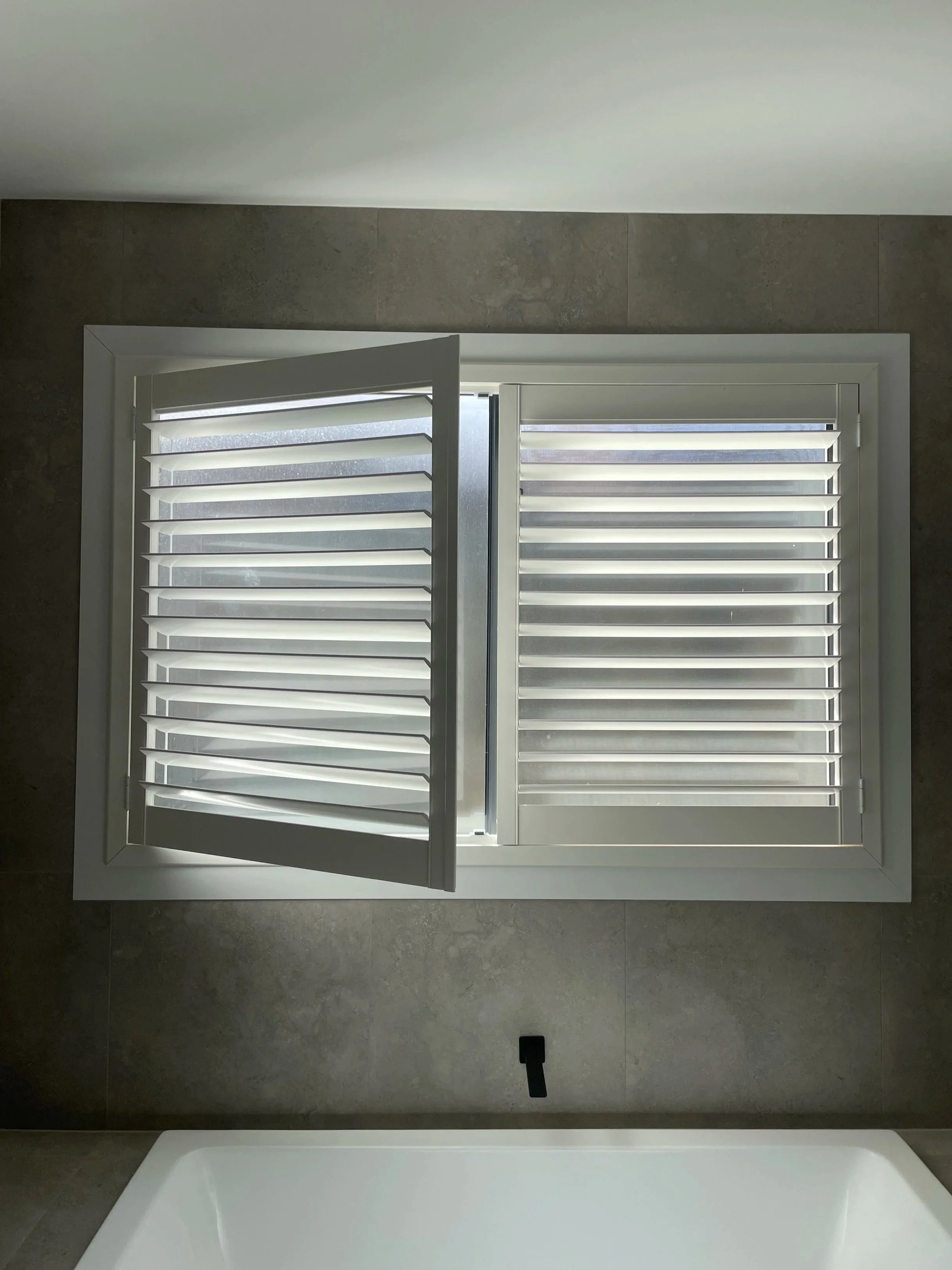 Bathroom window with white plantation shutters, partly open, above a bathtub with a faucet.