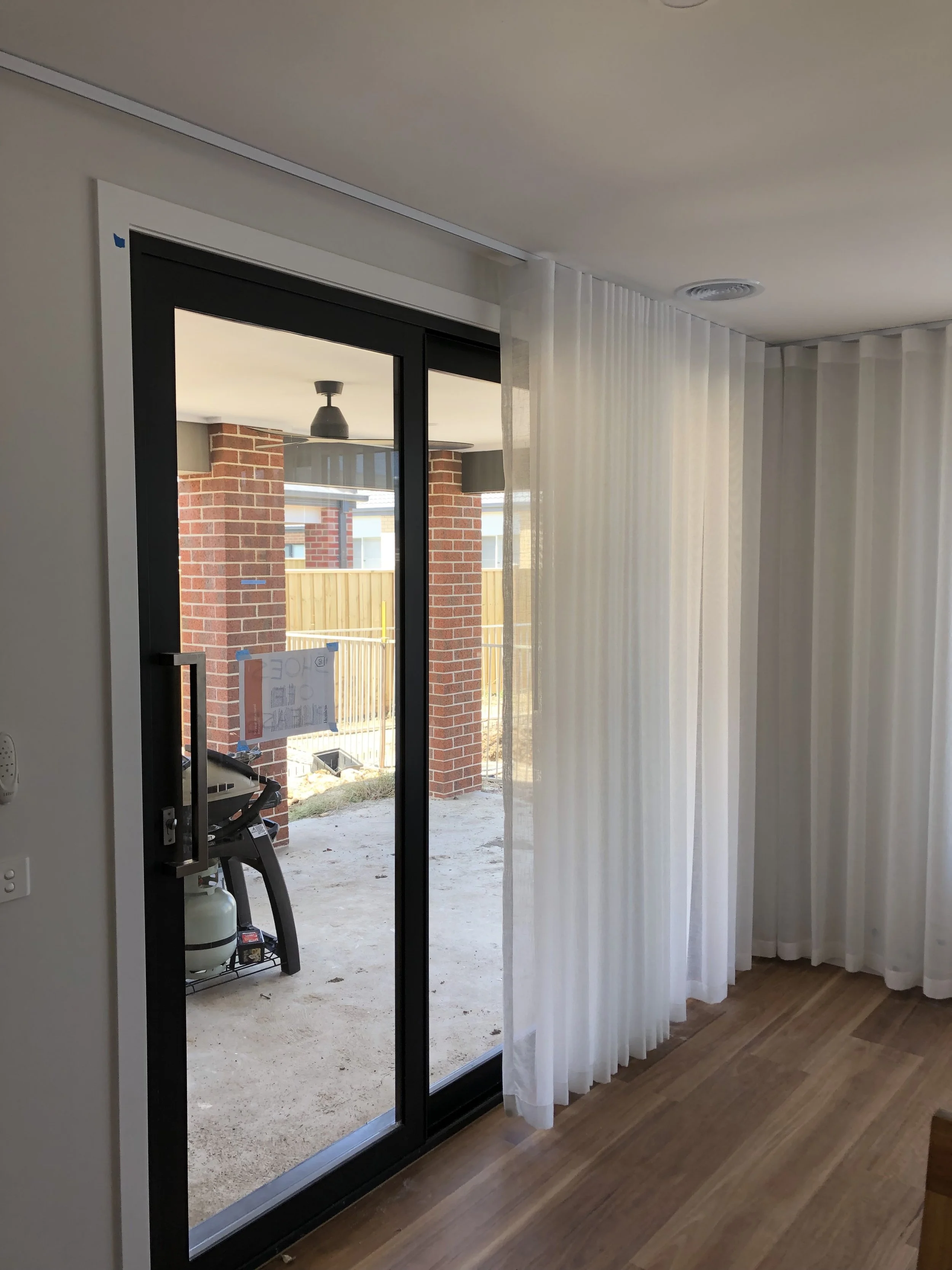 Interior view of a room with floor-to-ceiling white curtains and a sliding glass door leading to an outdoor patio with a barbecue grill and a brick column in the background.