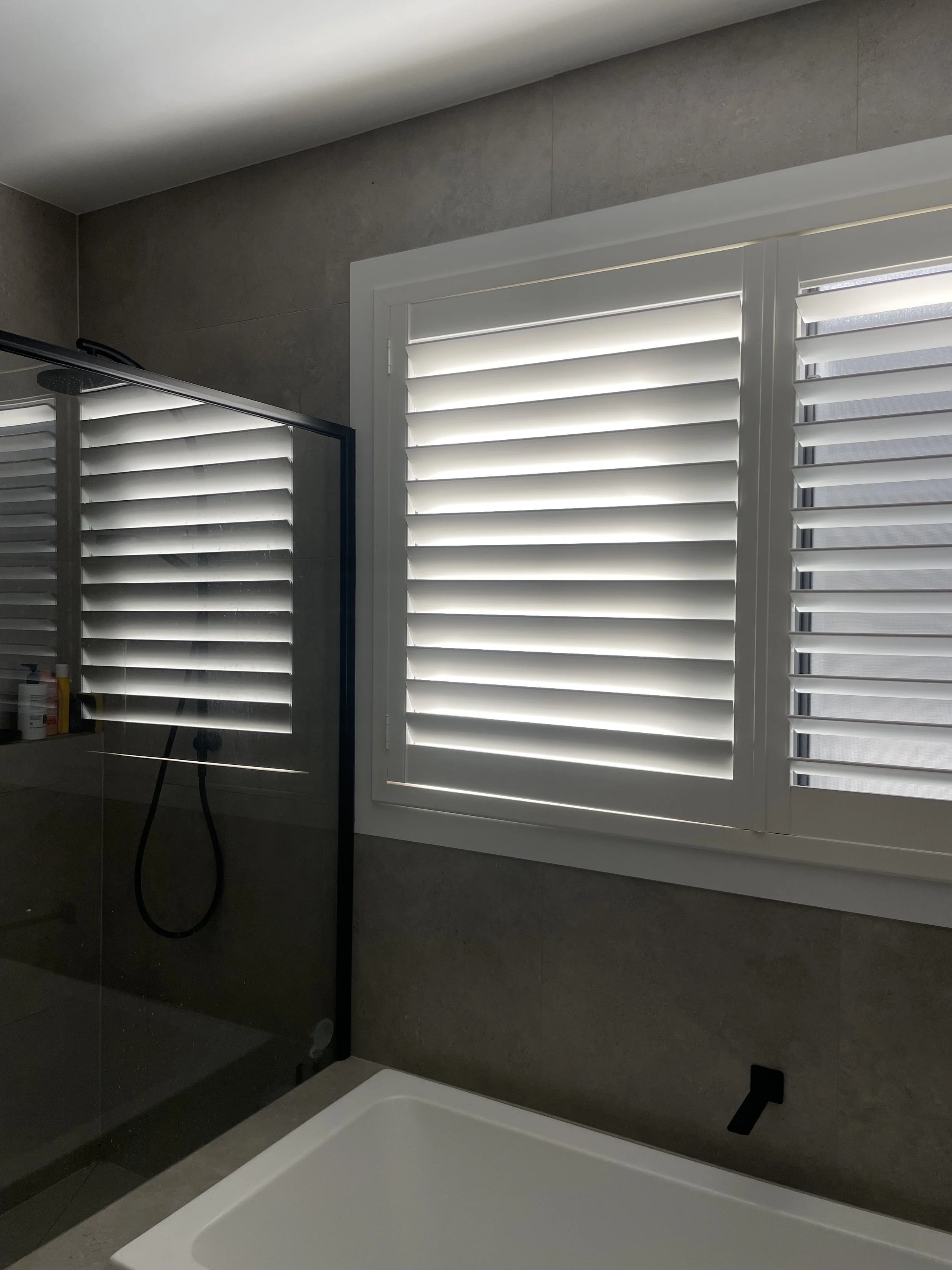 Modern bathroom with gray tile walls, white bathtub, a large window with white plantation shutters, and a glass shower enclosure with a black frame.