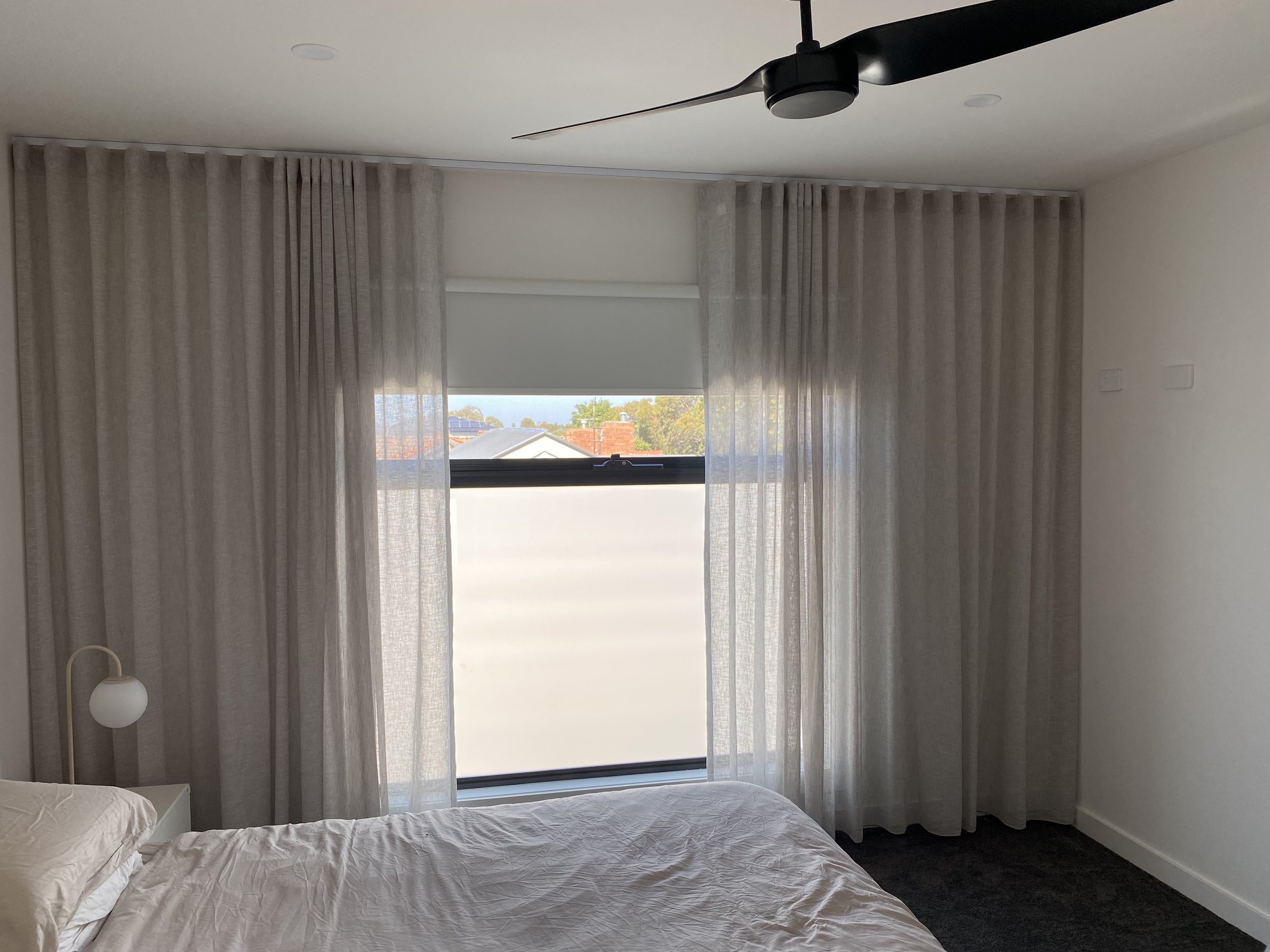 A bedroom with beige curtains, a bed with beige sheets, a white nightstand with a white spherical lamp, a window with a frosted lower glass pane and a view of rooftops and trees outside, a ceiling fan, and white walls.