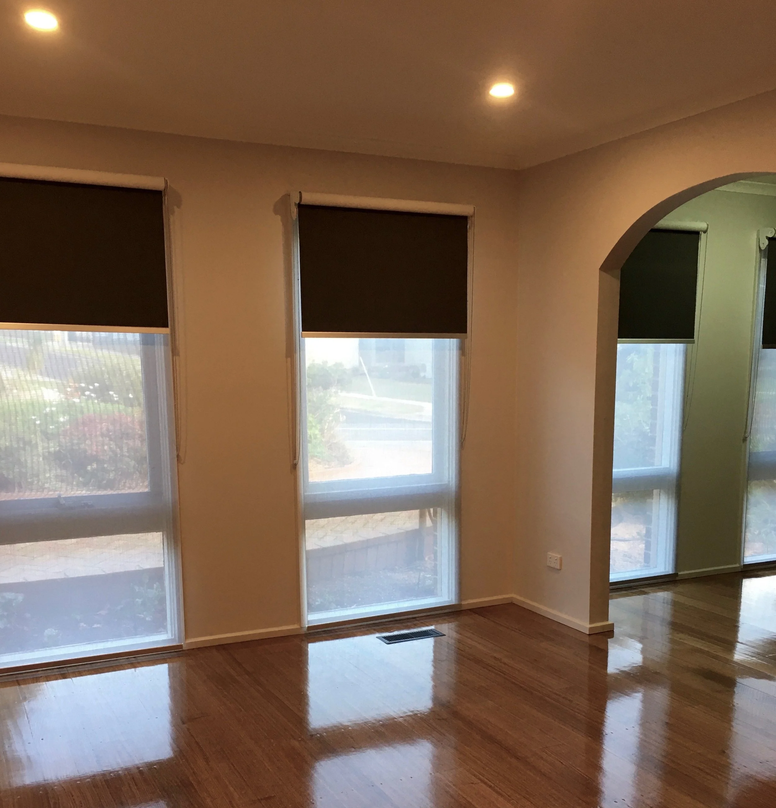 Empty living room with hardwood floors, three large windows with black roller shades, and an archway leading to another room. Recessed ceiling light is on.