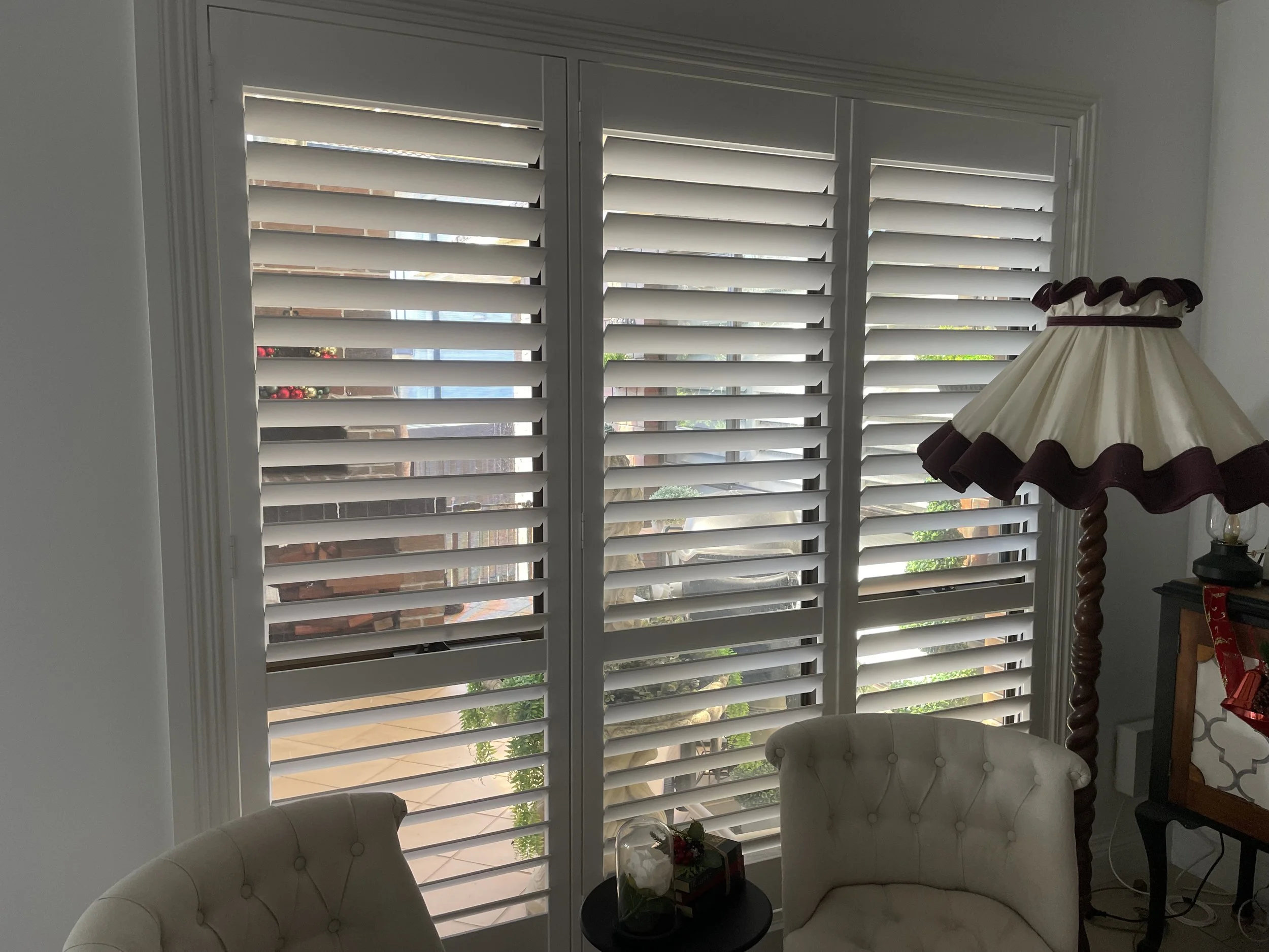 Living room with a large window covered by white plantation shutters, a vintage-style floor lamp with a cream and maroon shade, two cream upholstered chairs, and a small round black side table with a glass terrarium and Christmas decorations.