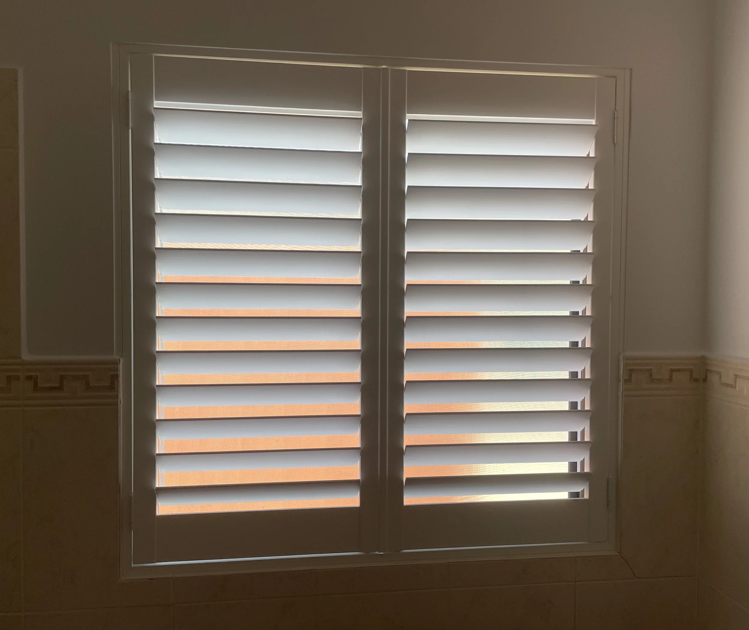 A window with white plantation shutters in a room with beige tiled walls.