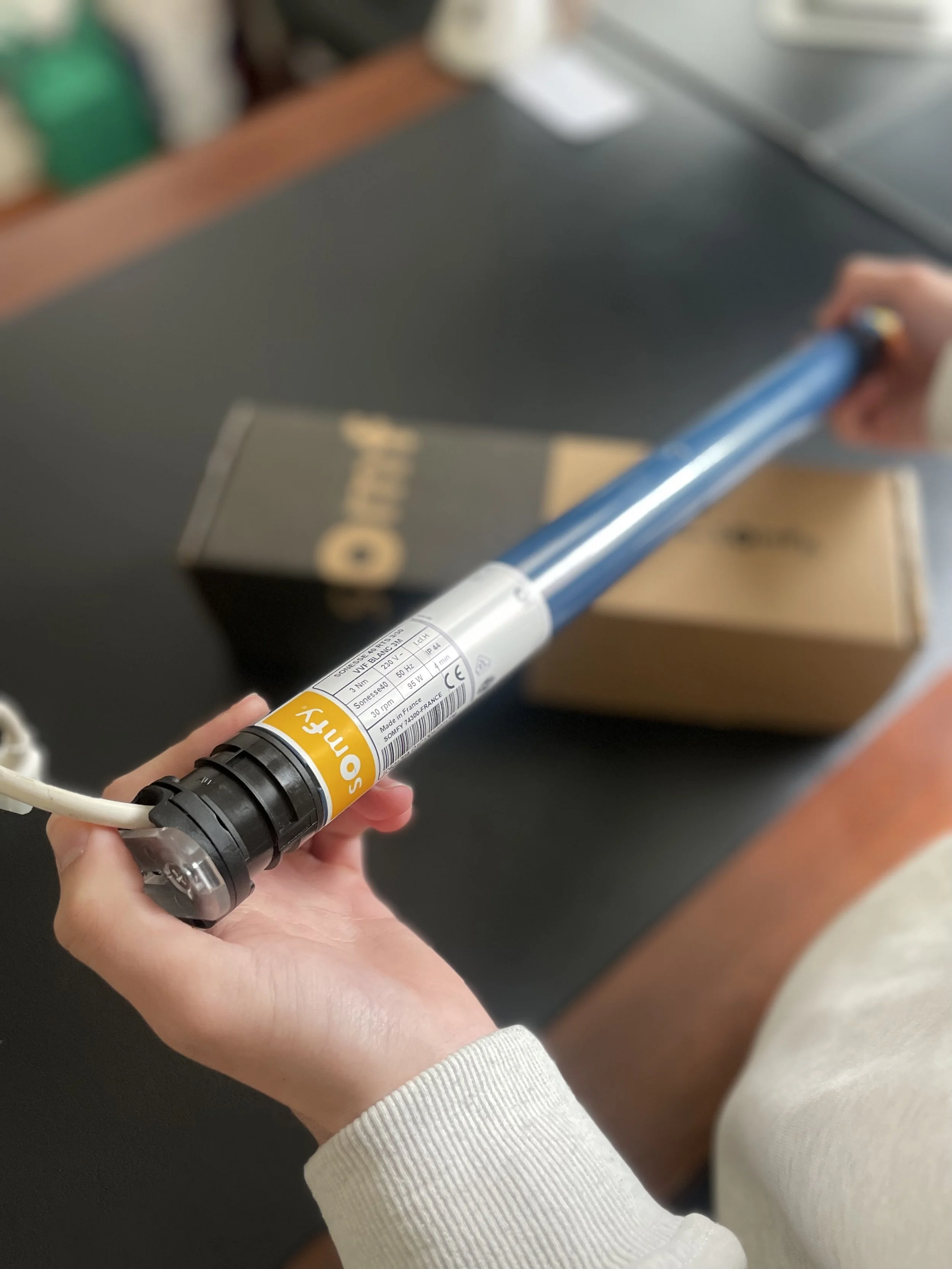 Hand holding a blue and white electric appliance with a visible orange and white label, against a blurred background of a desk with a box and other objects.