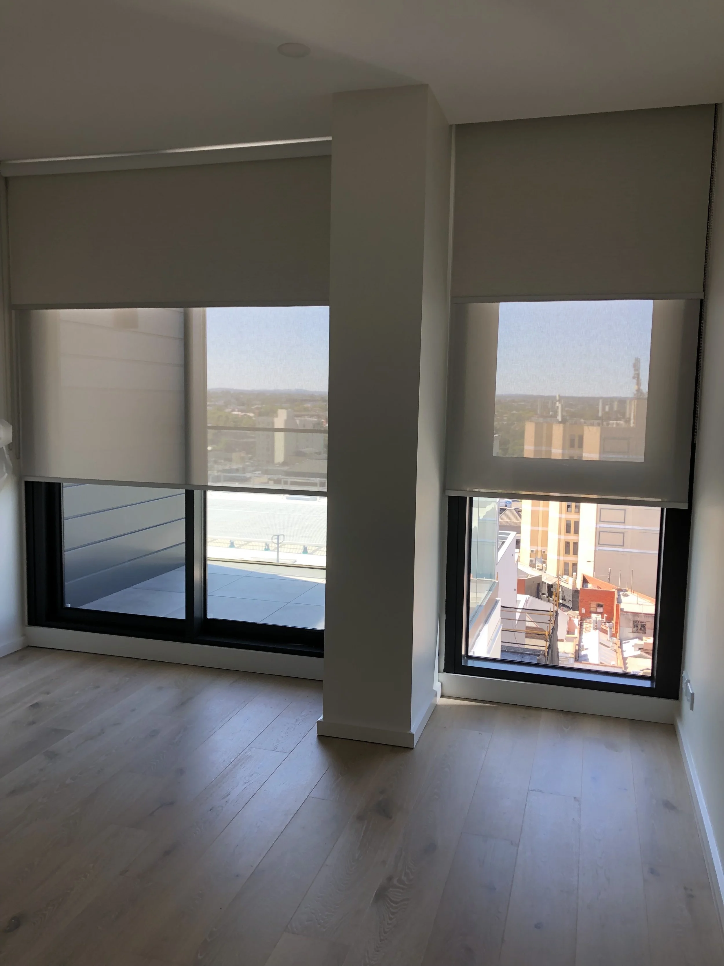 Empty room with two large windows with view of city buildings, partially covered by white roller blinds, and light wooden flooring.