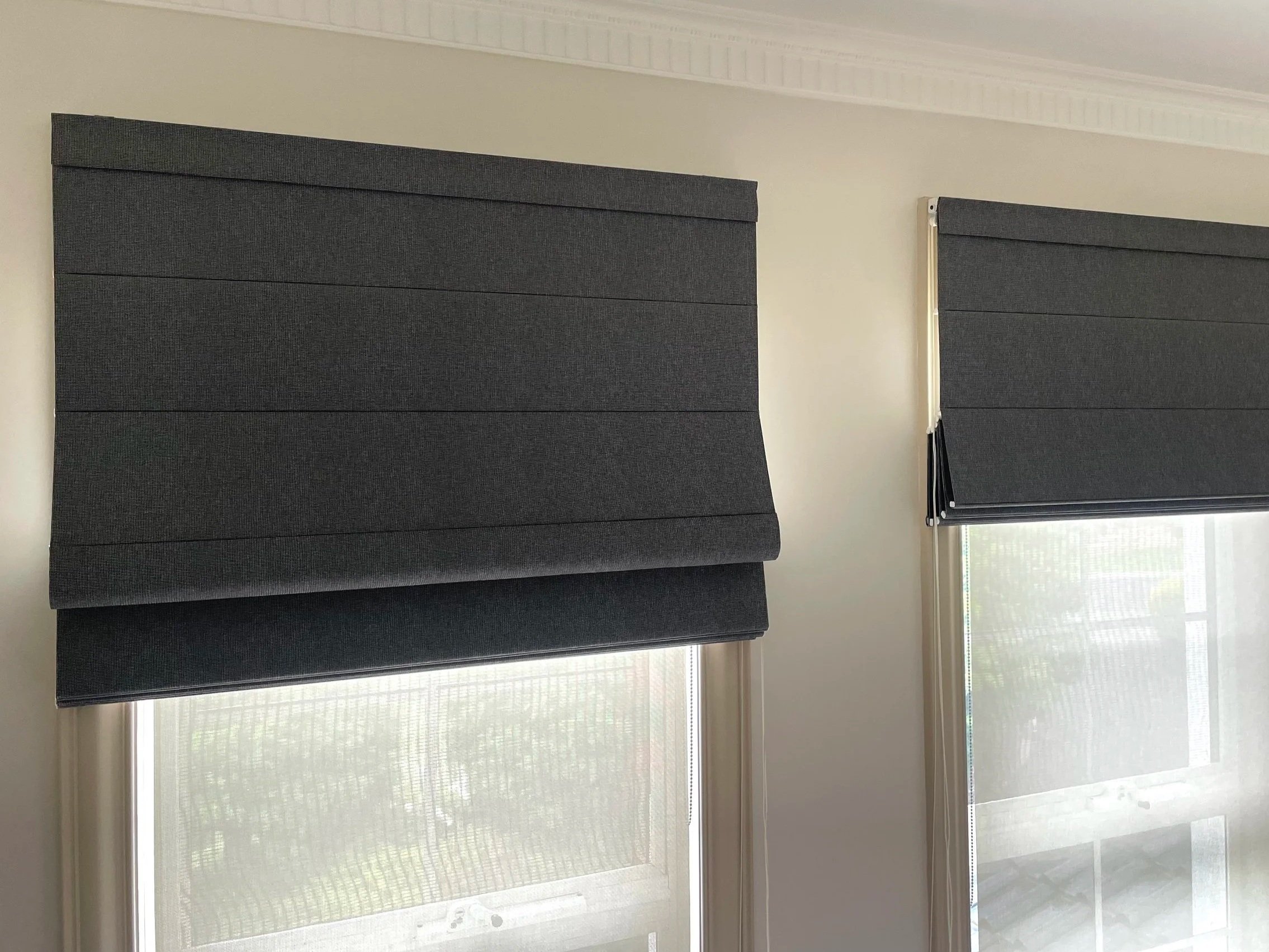 Two windows with dark gray Roman shades partially pulled down in a room with white walls and crown molding.