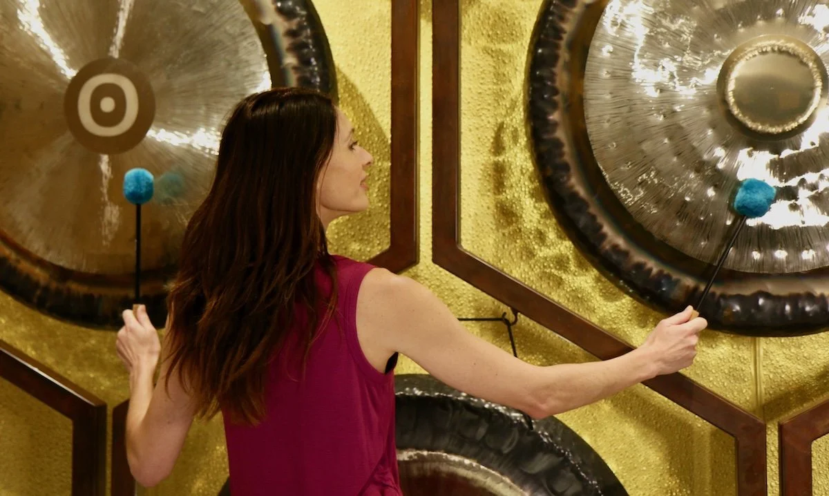A woman wearing a pink sleeveless top strikes a gong with a mallet with a blue head.